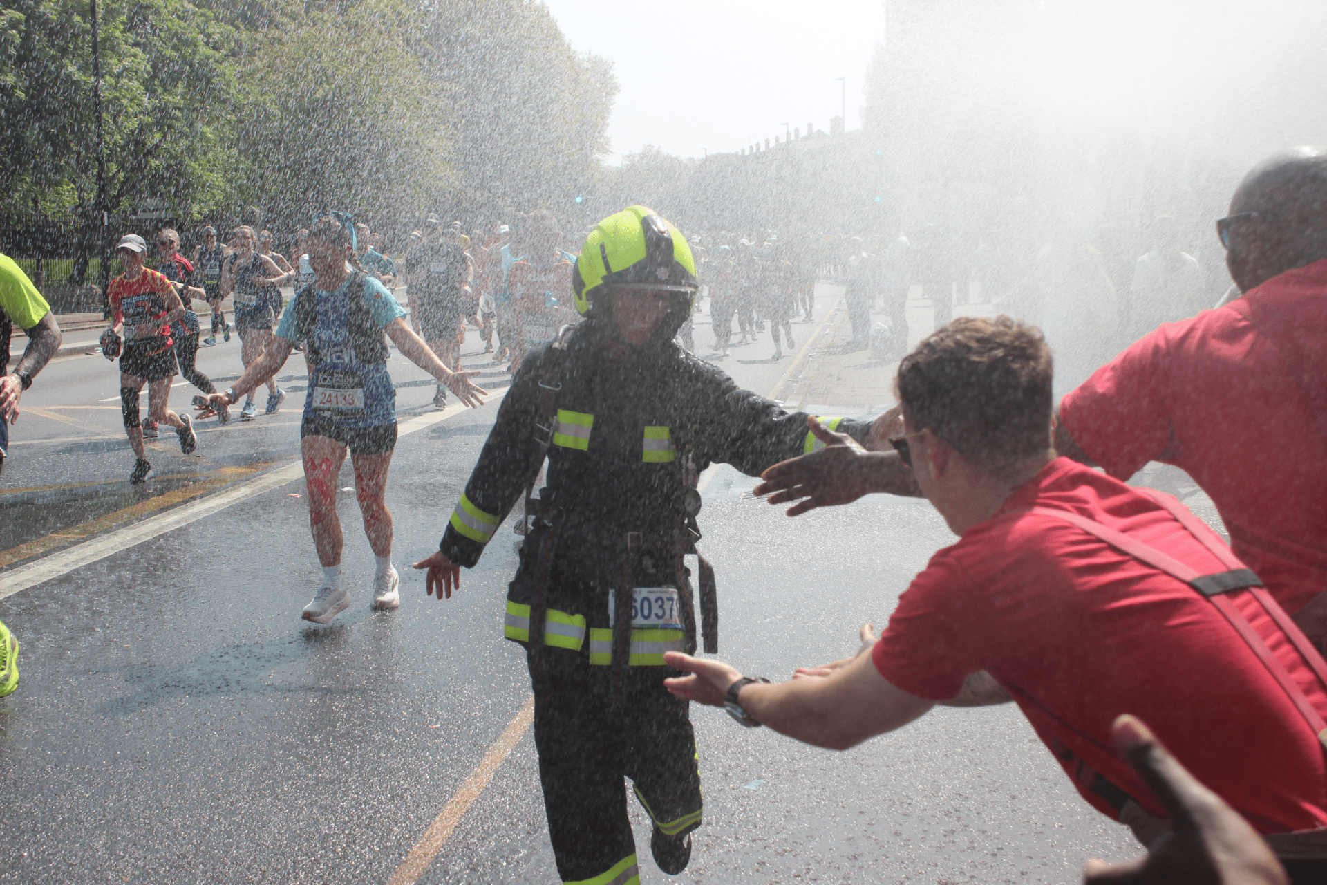 A firefighter in full gear runs through mist from a hose, high-fiving a supporter as marathon runners pass by on a sunny street.