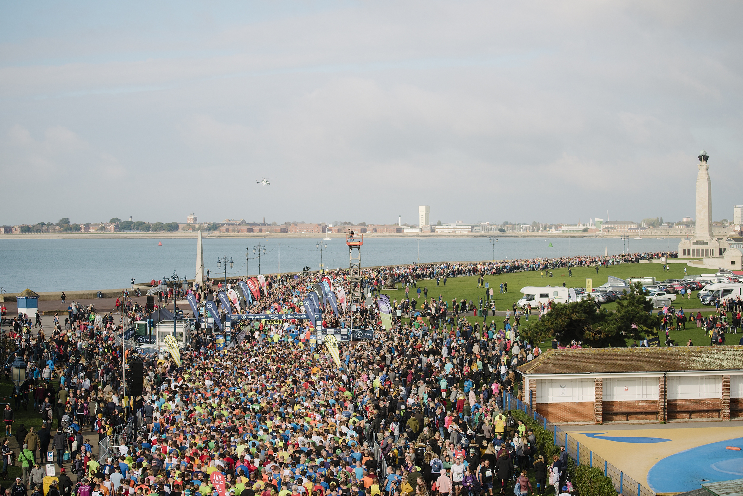 A large crowd of marathon runners gathers at the starting line near a waterfront park, with spectators lining both sides under a partly cloudy sky. Buildings, water, and a tall monument are visible in the background.