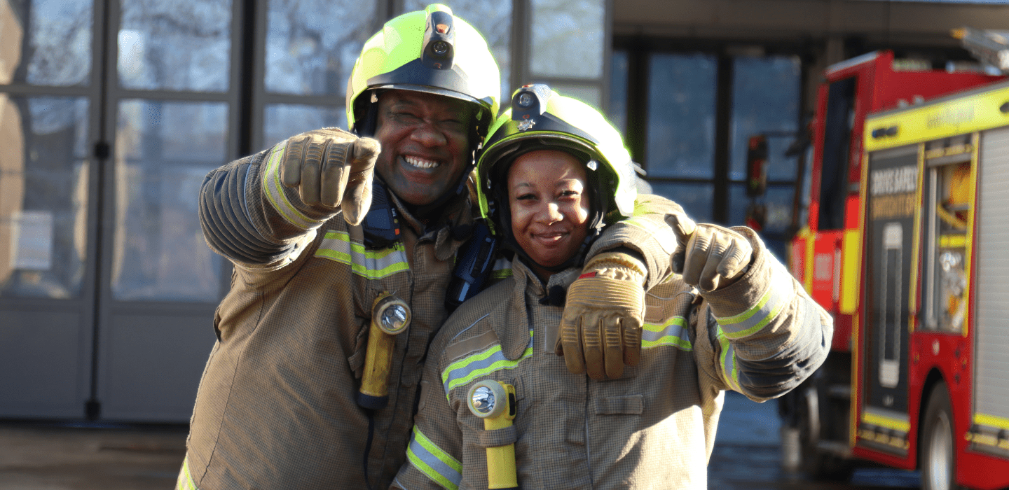 Two firefighters in protective gear smile and point toward the camera, standing close together in front of a fire truck and building.