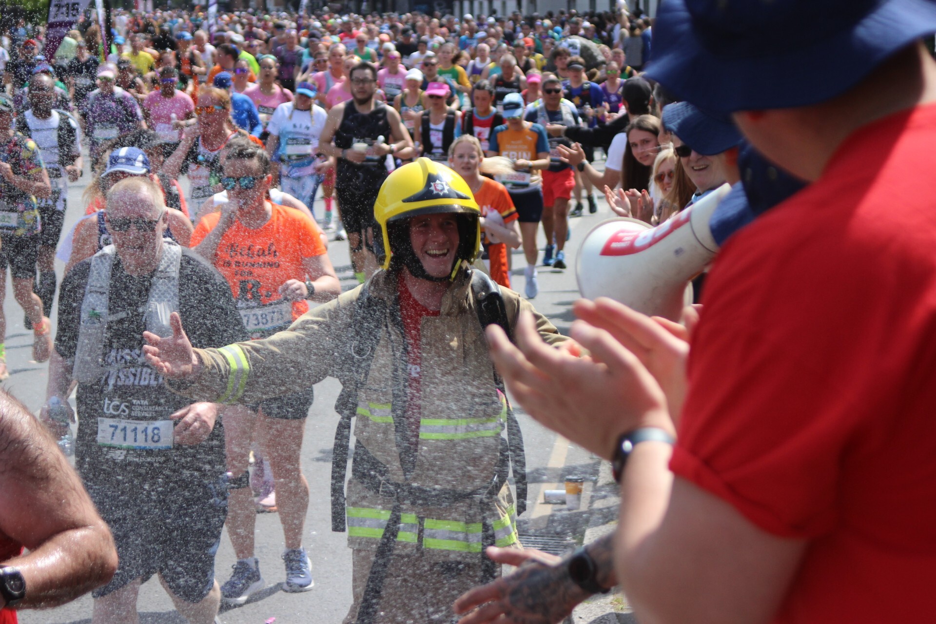 A firefighter in full gear smiles while running in a crowded marathon, as people on the sidelines cheer and spray water, creating a festive, spirited atmosphere.