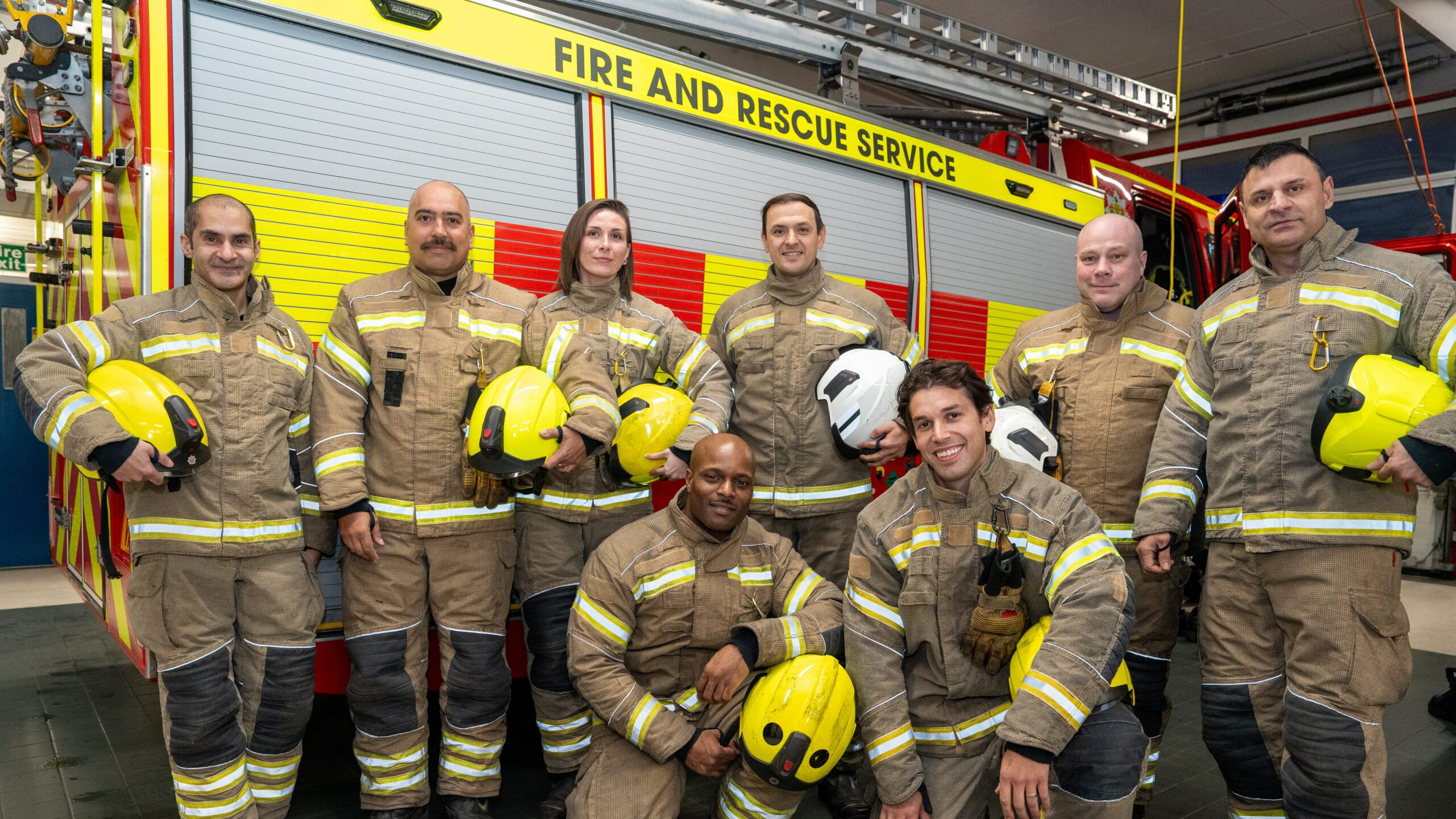 Eight firefighters in uniform pose in front of a fire truck labeled “Fire and Rescue Service.” Some stand, some kneel, all holding helmets and smiling at the camera.