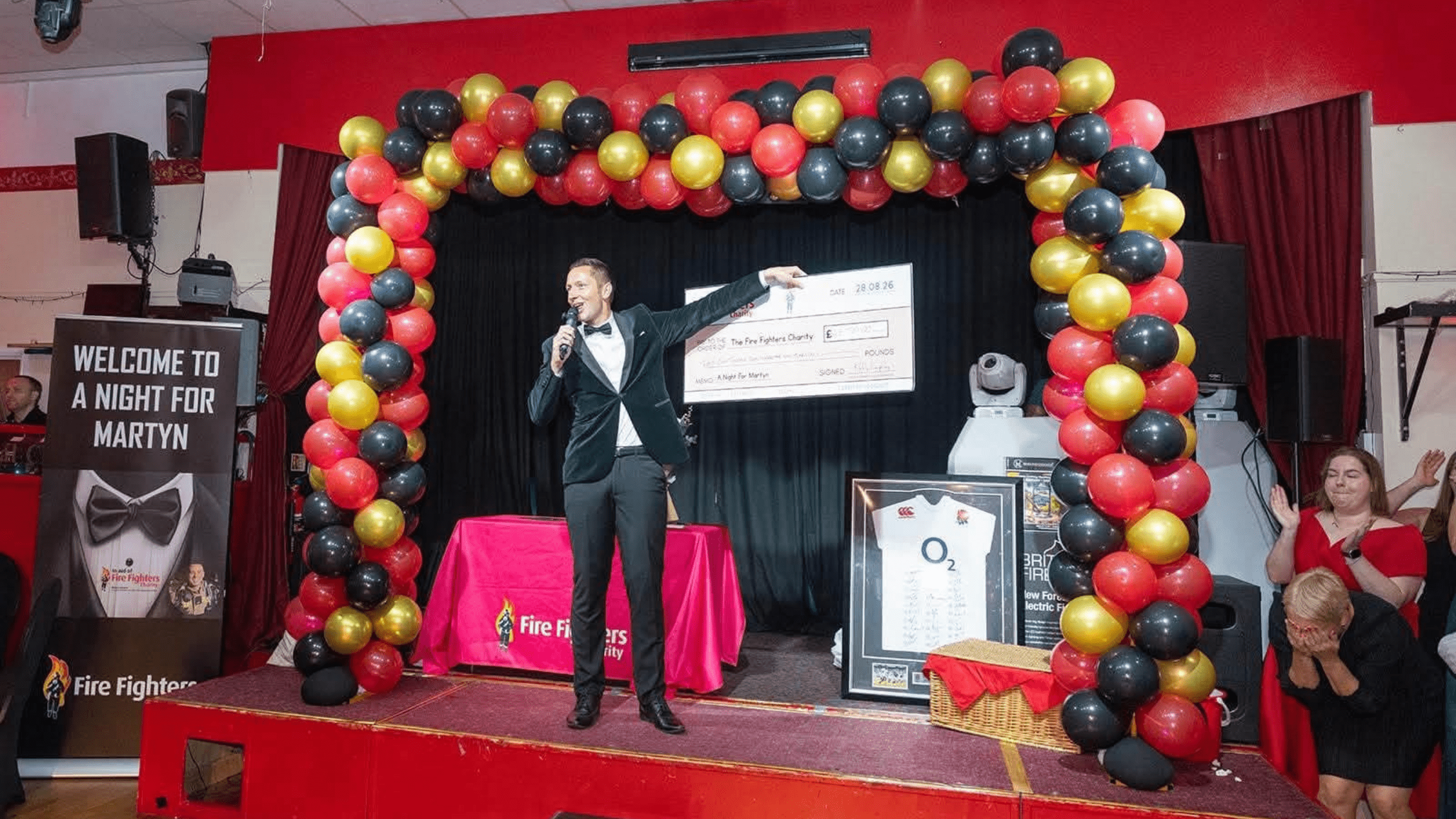 A man in a tuxedo holds a large check on stage under a red, black, and gold balloon arch at an event. A sign reads “Welcome to a Night for Martyn.” People applaud in the background.