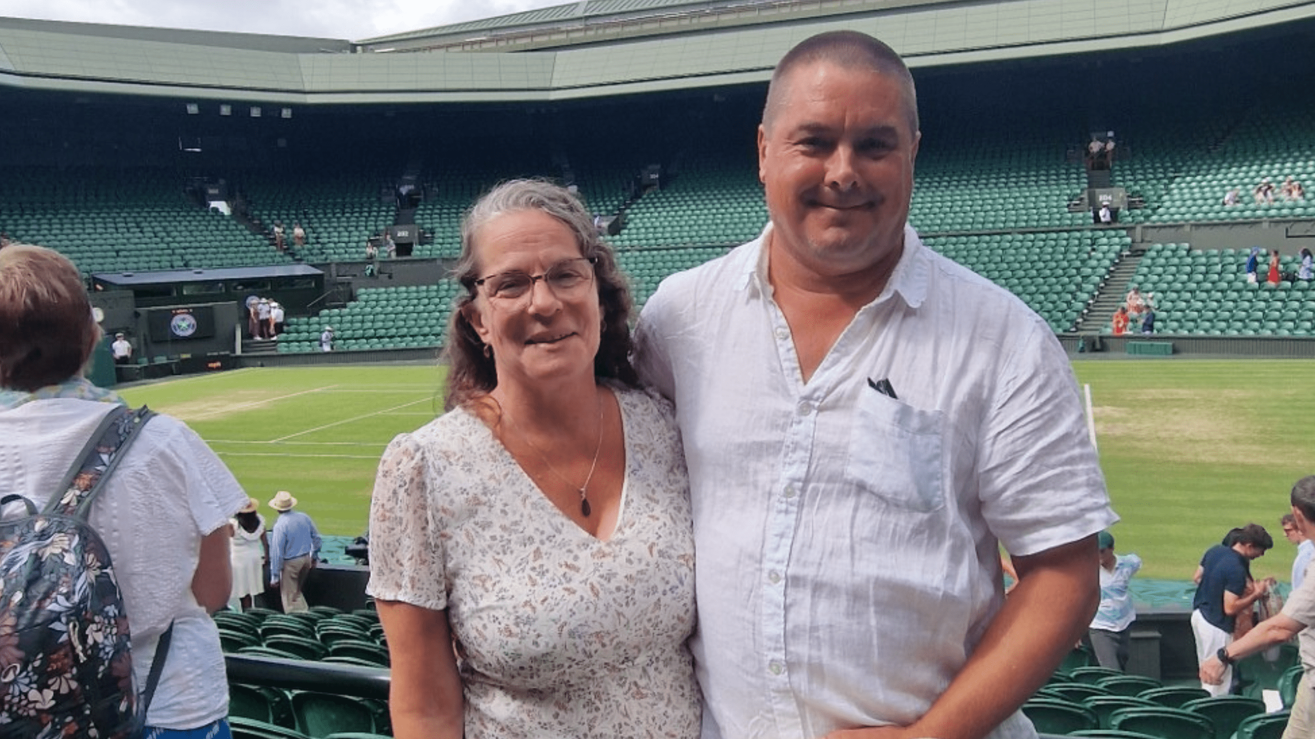 A smiling man and woman stand together at a tennis stadium, with empty green seats behind them. The man wears a white shirt and the woman wears a floral top. The grass tennis court is visible in the background.