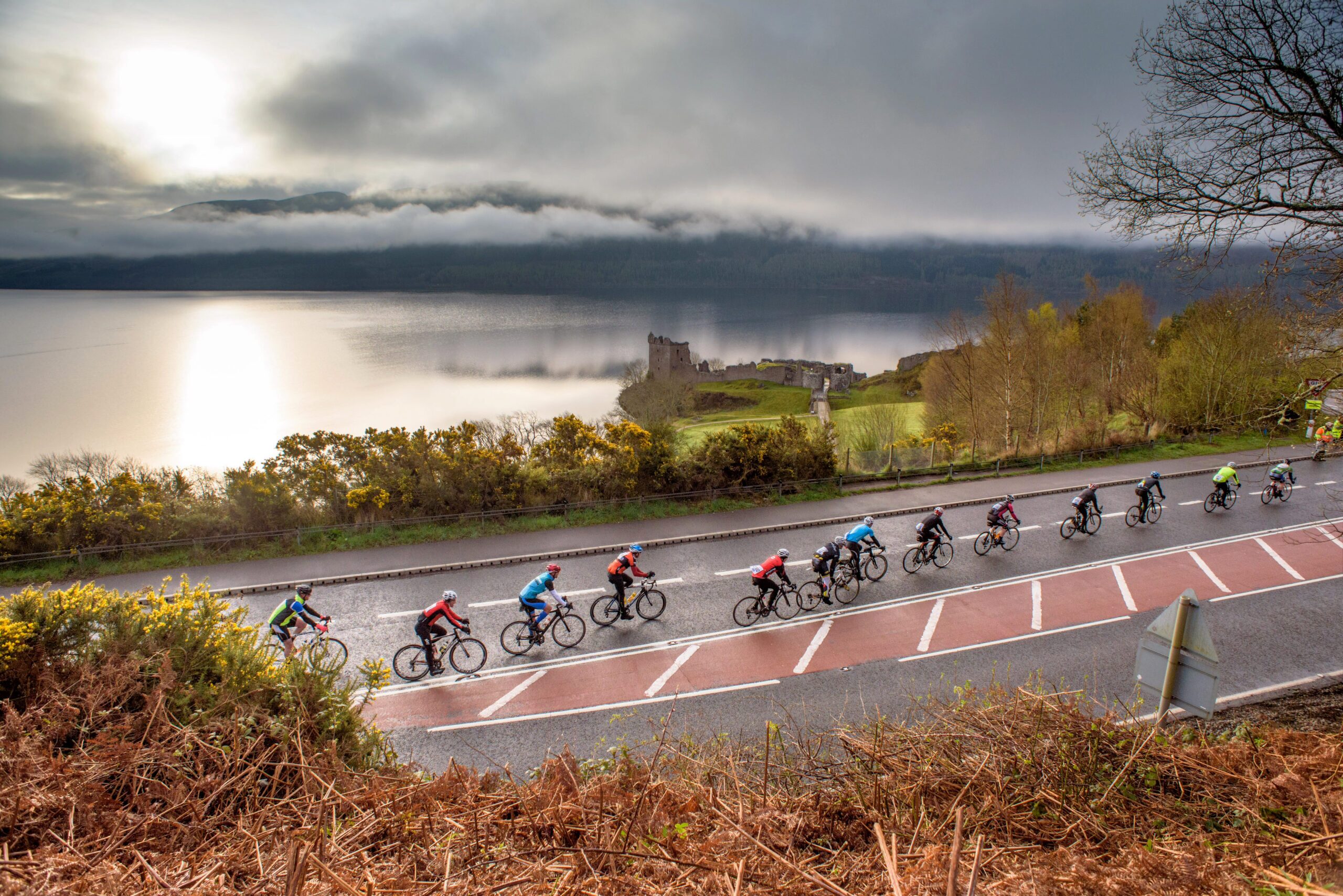 A group of cyclists rides in a single file line along a road bordering Loch Ness, with a castle ruin visible in the distance during the Etape Loch Ness sportive.