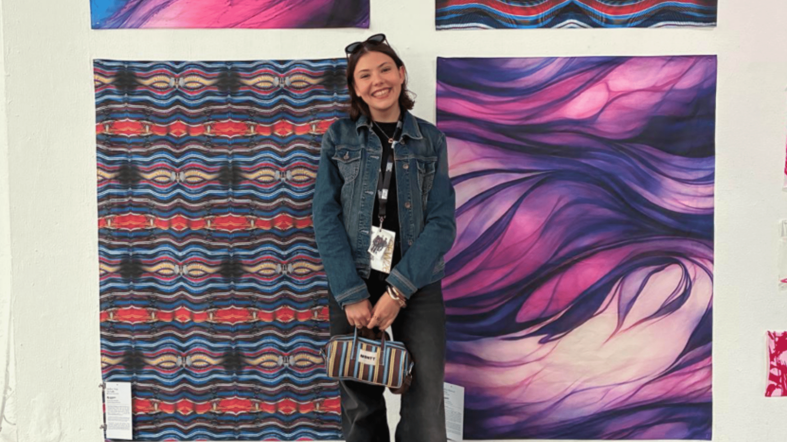 A woman in a denim jacket smiles and stands in front of two colorful abstract artworks—one with wavy purple patterns and one with red, blue, and white zigzags—displayed on a white wall. She holds a striped bag.