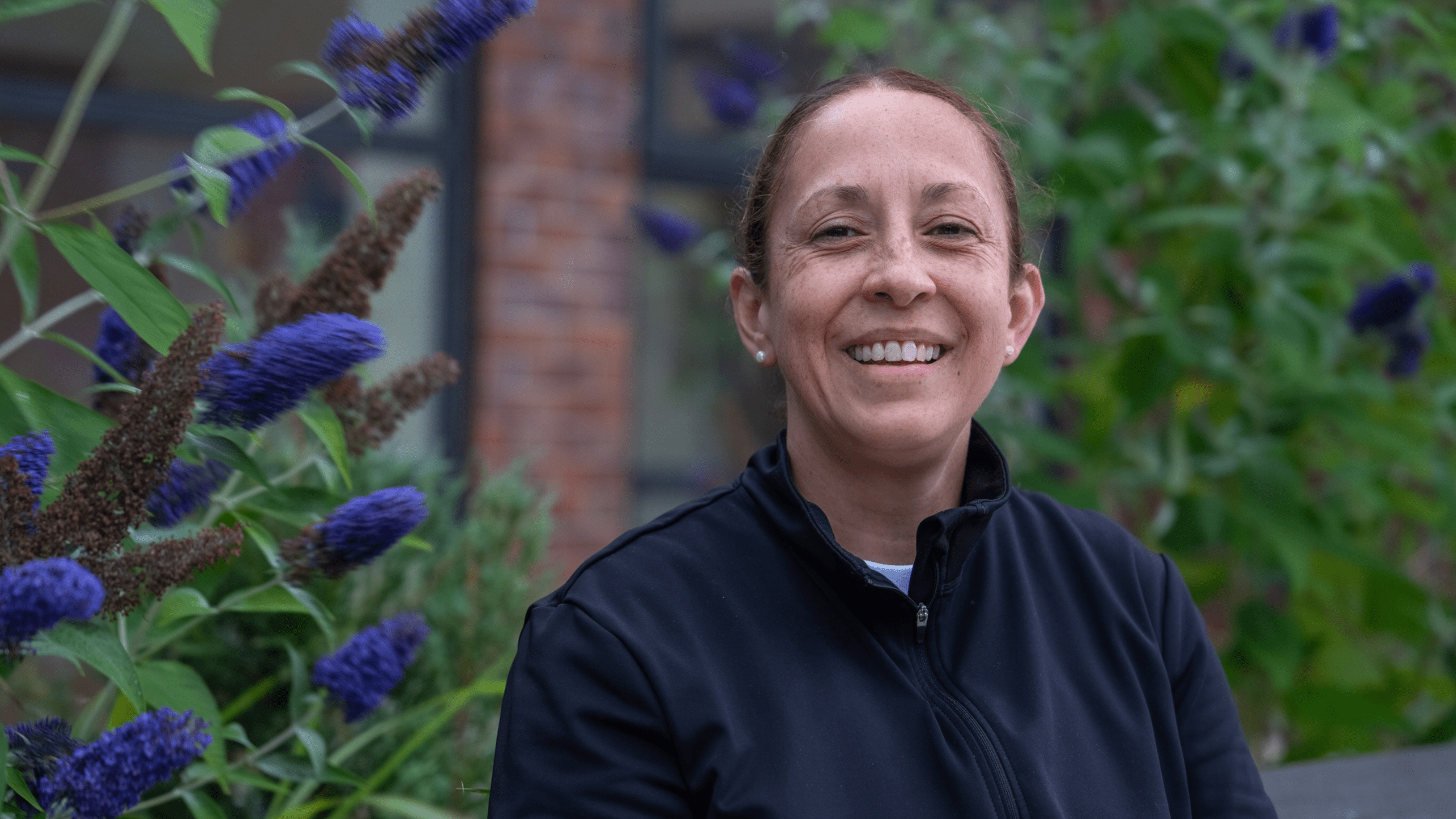 A smiling woman in a black zip-up jacket posing in front of a blooming butterfly bush.
