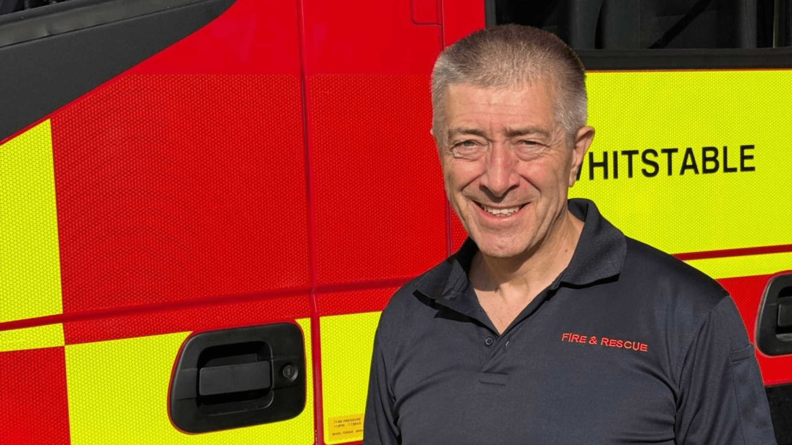 A smiling man wearing a Fire & Rescue polo shirt stands in front of a red and yellow fire engine labeled “Whitstable.”