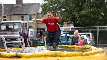 A smiling man in firefighting gear falls into a pool of water from a seat while people, including children, watch and cheer near parked vehicles and buildings outdoors.