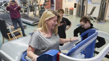 A woman with blonde hair smiles while using a specialized treadmill in a rehabilitation facility. Two Fire Fighters Charity staff stand nearby, watching and guiding her.