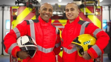 Two firefighters in bright red uniforms stand smiling, holding helmets. They are in front of a fire truck with chevron stripes inside a garage. One holds a white helmet, the other a yellow one.