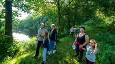 A group of people, including adults and children, walk together along a grassy, tree-lined path next to a lake near Harcombe House on a sunny day. Sunlight filters through the leafy trees, creating a bright and peaceful atmosphere.
