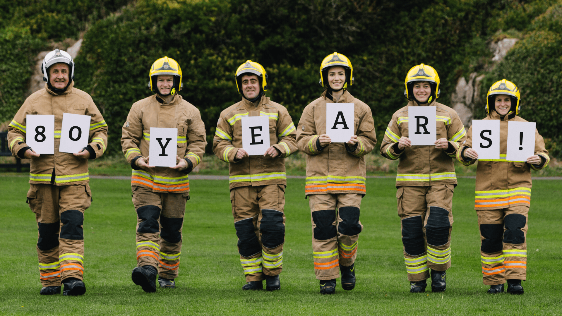 Six firefighters in full gear stand on grass, each holding a sign. Together, the signs read 