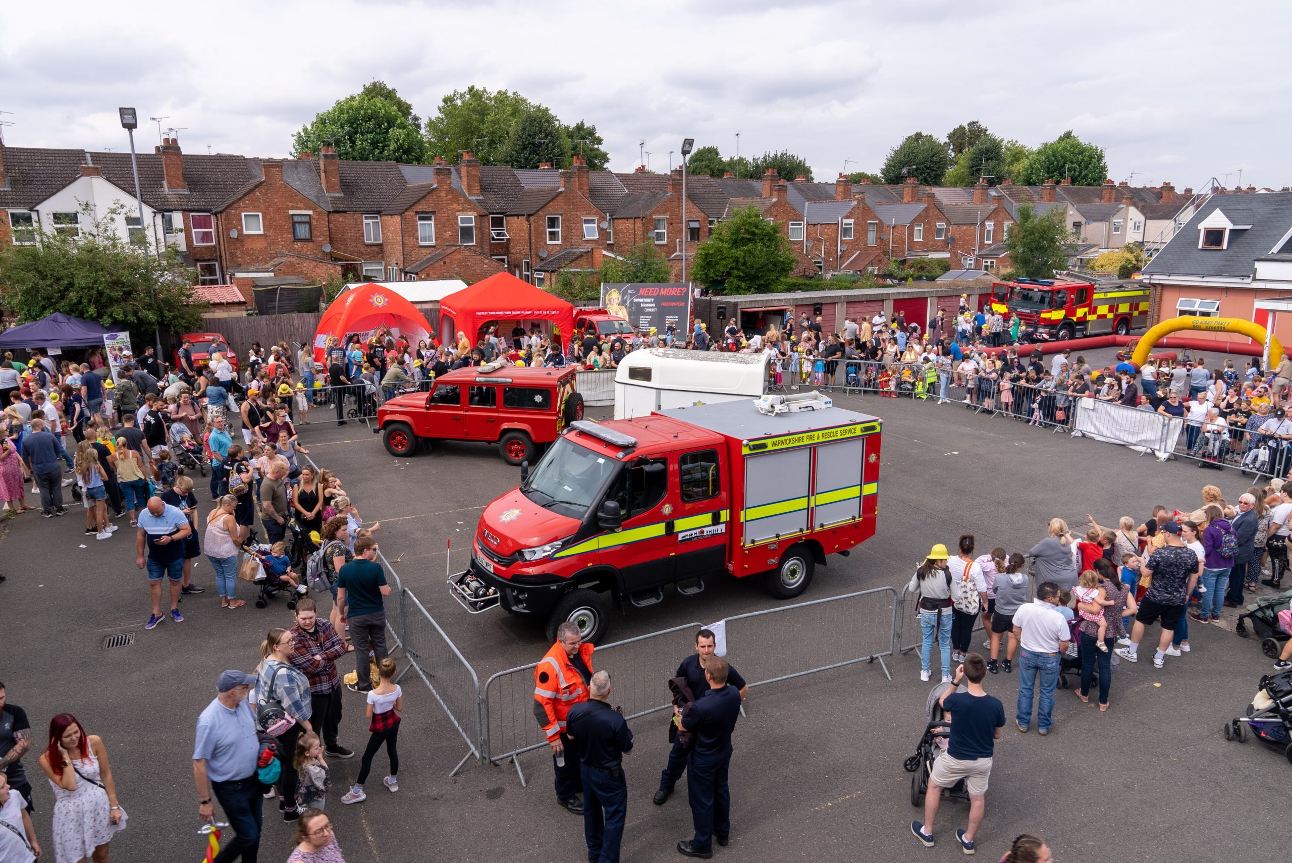 A busy outdoor fire station community open day features crowds of people, two fire trucks, tents, and barriers in a parking lot with houses in the background on a cloudy day. Emergency service personnel are present among attendees.
