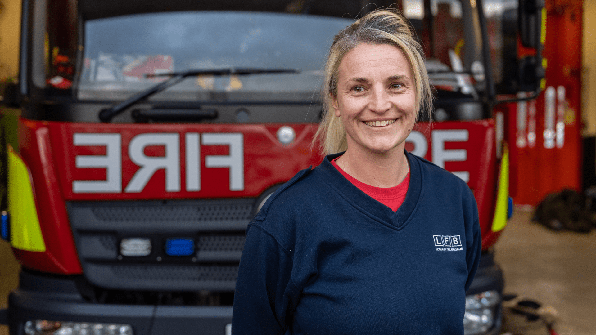 A smiling firefighter stands in front of a fire engine inside a fire station, wearing a navy blue uniform. The word 