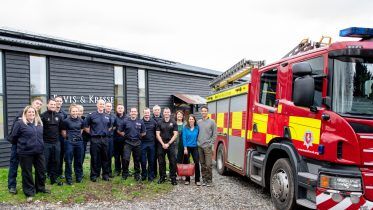 A group of people, including firefighters in uniform and civilians, stand outside the Elvis and Kresse workshop with a fire engine parked outside.