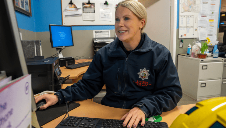 A woman in a Cumbria Fire & Rescue Service jacket sits at a desk, using a computer. She is smiling, with office equipment, documents, and bulletin boards in the background.