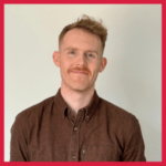 A man with short red hair and a mustache, wearing a buttoned-up brown shirt, stands against a plain off-white background. The image has a red border.