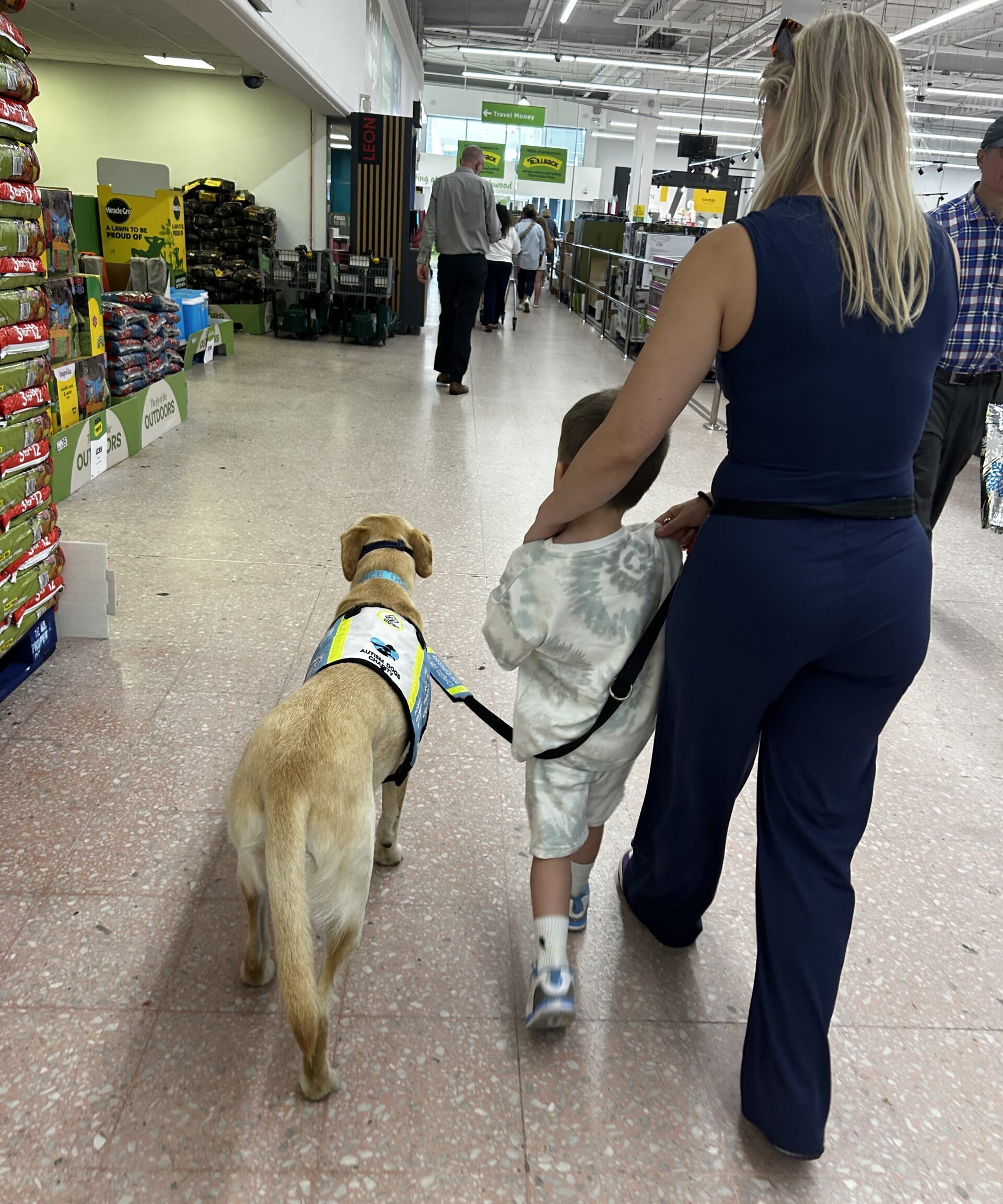 A woman and a child walk through a store, with the child holding a leash connected to a yellow Labrador guide dog wearing a yellow and blue vest. Shelves with pet supplies are visible on the left.