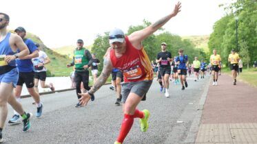 A smiling marathon runner in a red vest and neon shoes poses with arms outstretched toward the camera while other runners continue along a road bordered by greenery.