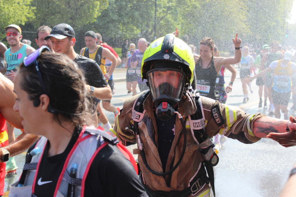 A firefighter in full gear and helmet runs a marathon among other runners, with water spraying in the background on a sunny day. Some runners are smiling, and one in the background gestures with raised middle fingers.