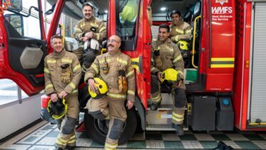 Five firefighters in uniform pose smiling by a red West Midlands Fire Service truck inside a fire station, holding yellow helmets and standing or sitting by the open vehicle doors.