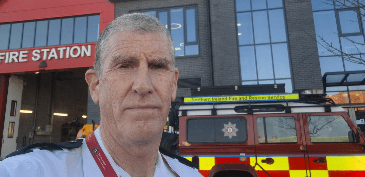 A man in a fire service uniform stands outside a fire station, with a red Northern Ireland Fire and Rescue Service vehicle parked behind him. The fire station building is visible in the background.