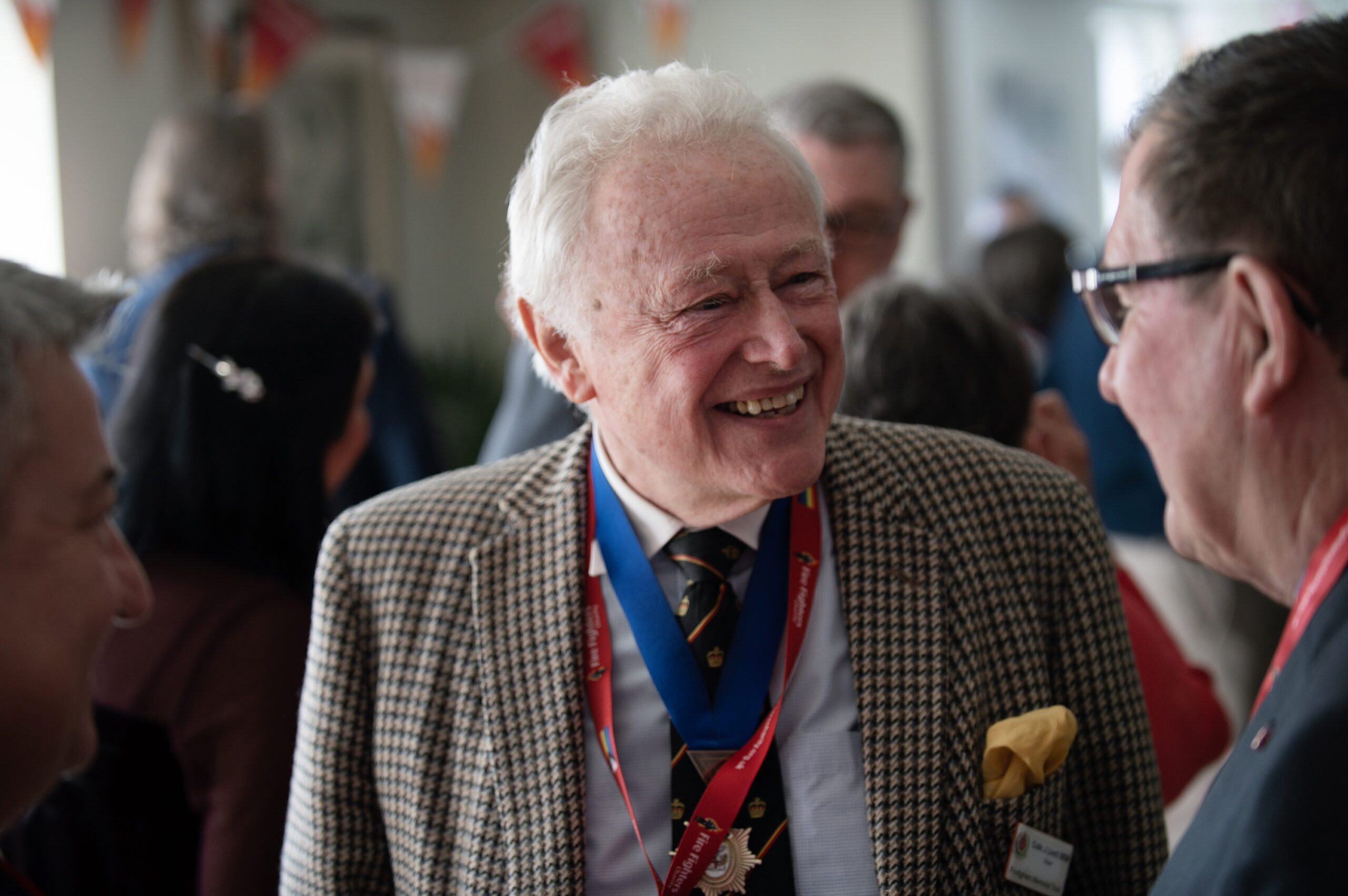 An elderly man in a checkered blazer and blue ribbon medal smiles and speaks with others at a social event. He is surrounded by people in a lively indoor setting.