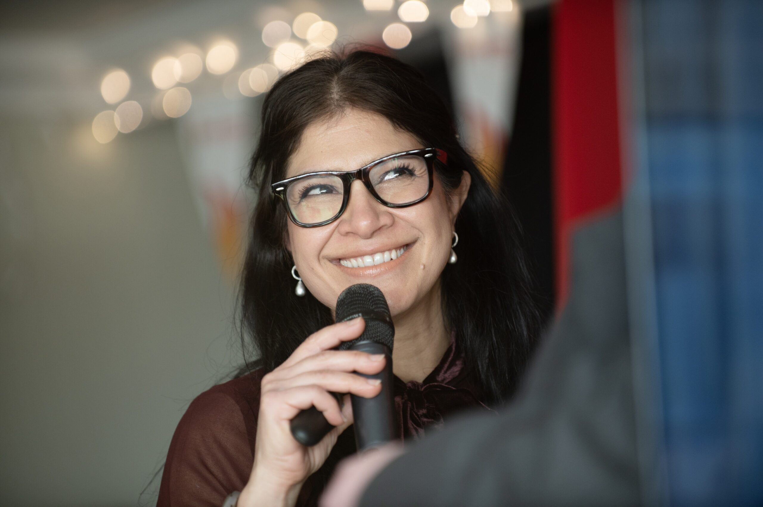 A woman with long dark hair and glasses smiles while holding a microphone. String lights and blurred decorations are visible in the background.