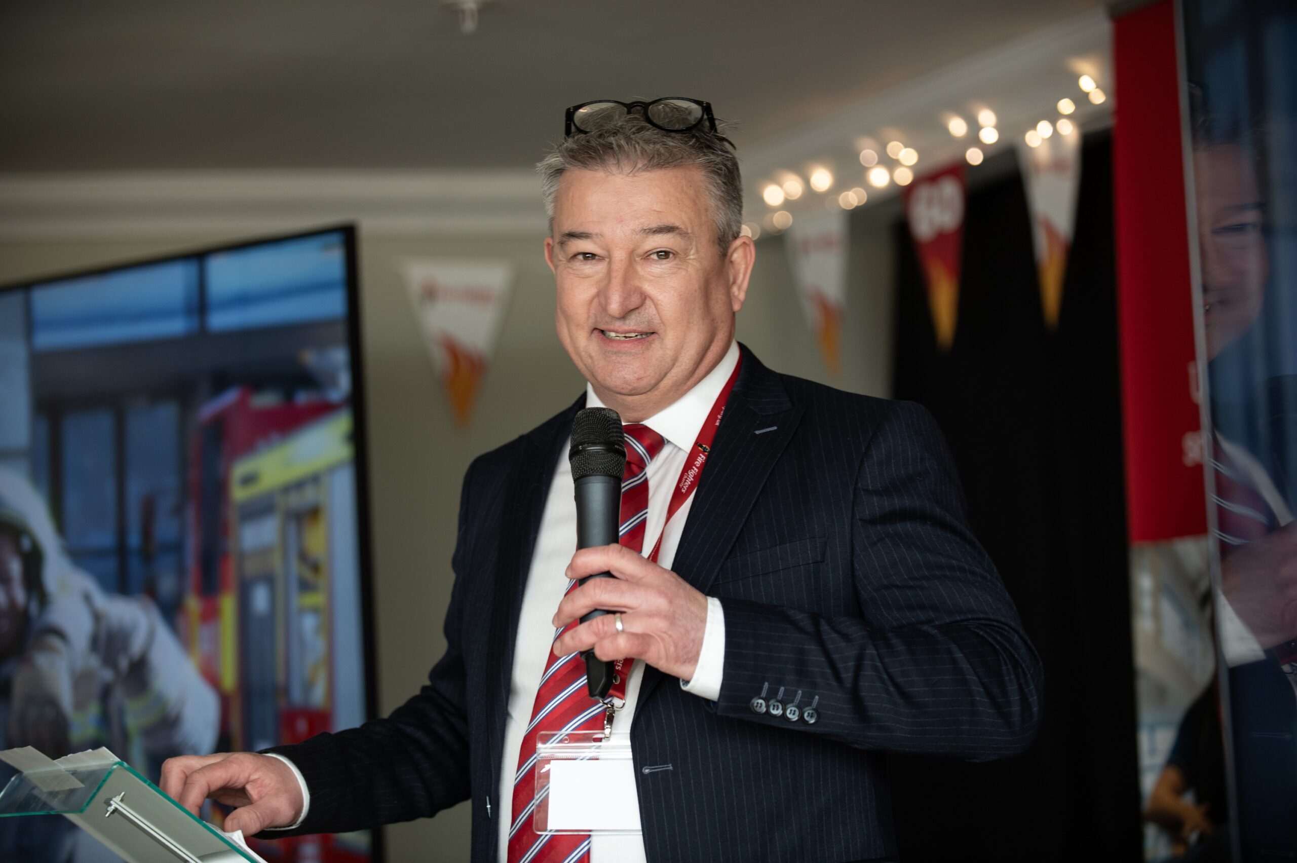 A man in a suit and red striped tie holds a microphone and speaks at an indoor event. He has glasses resting on his head and stands near a podium with a large screen and banners visible in the background.