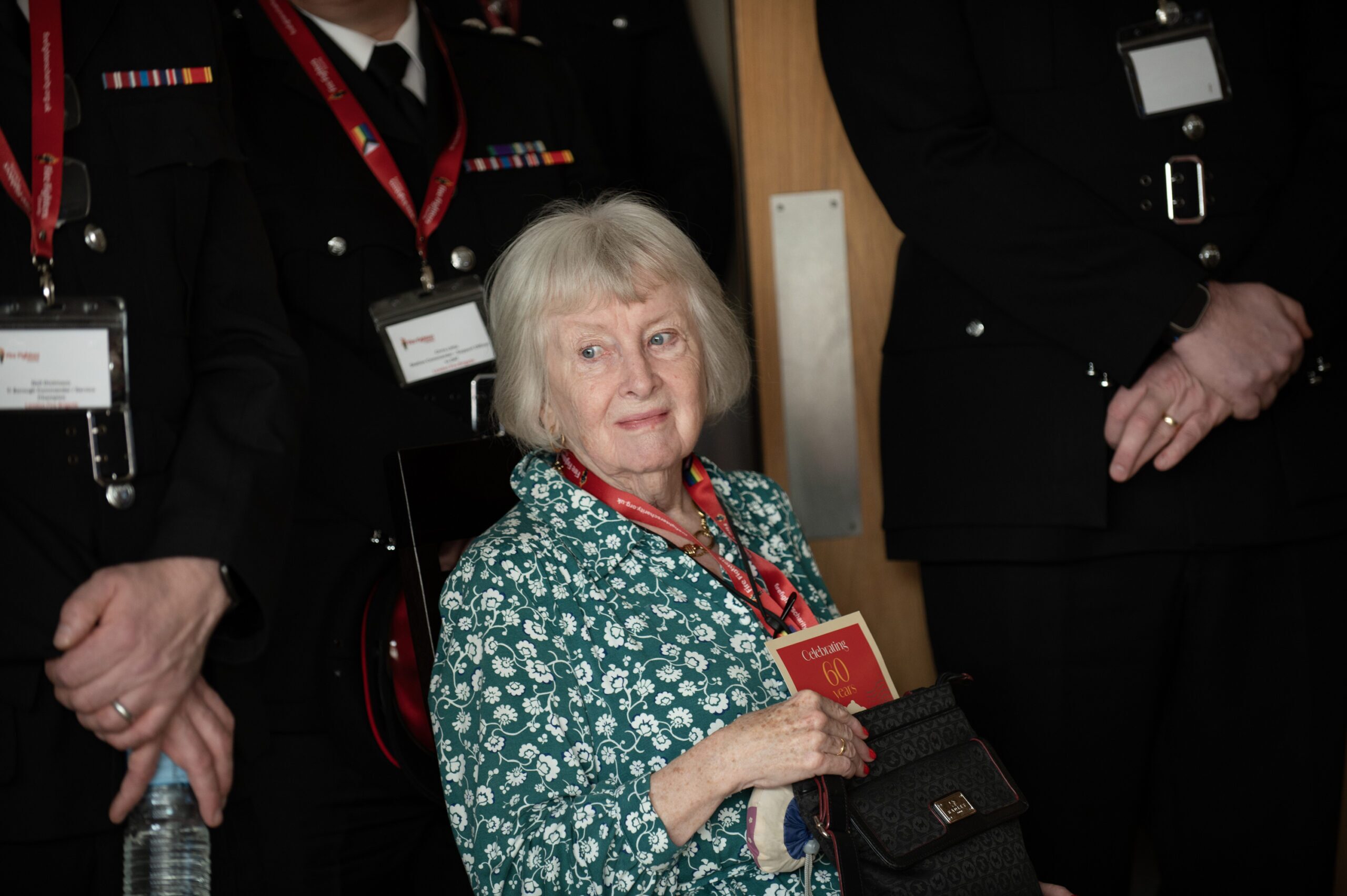 An older woman with white hair, wearing a green patterned top, sits holding a red book and a black purse, surrounded by people in black uniforms with red lanyards and name badges.