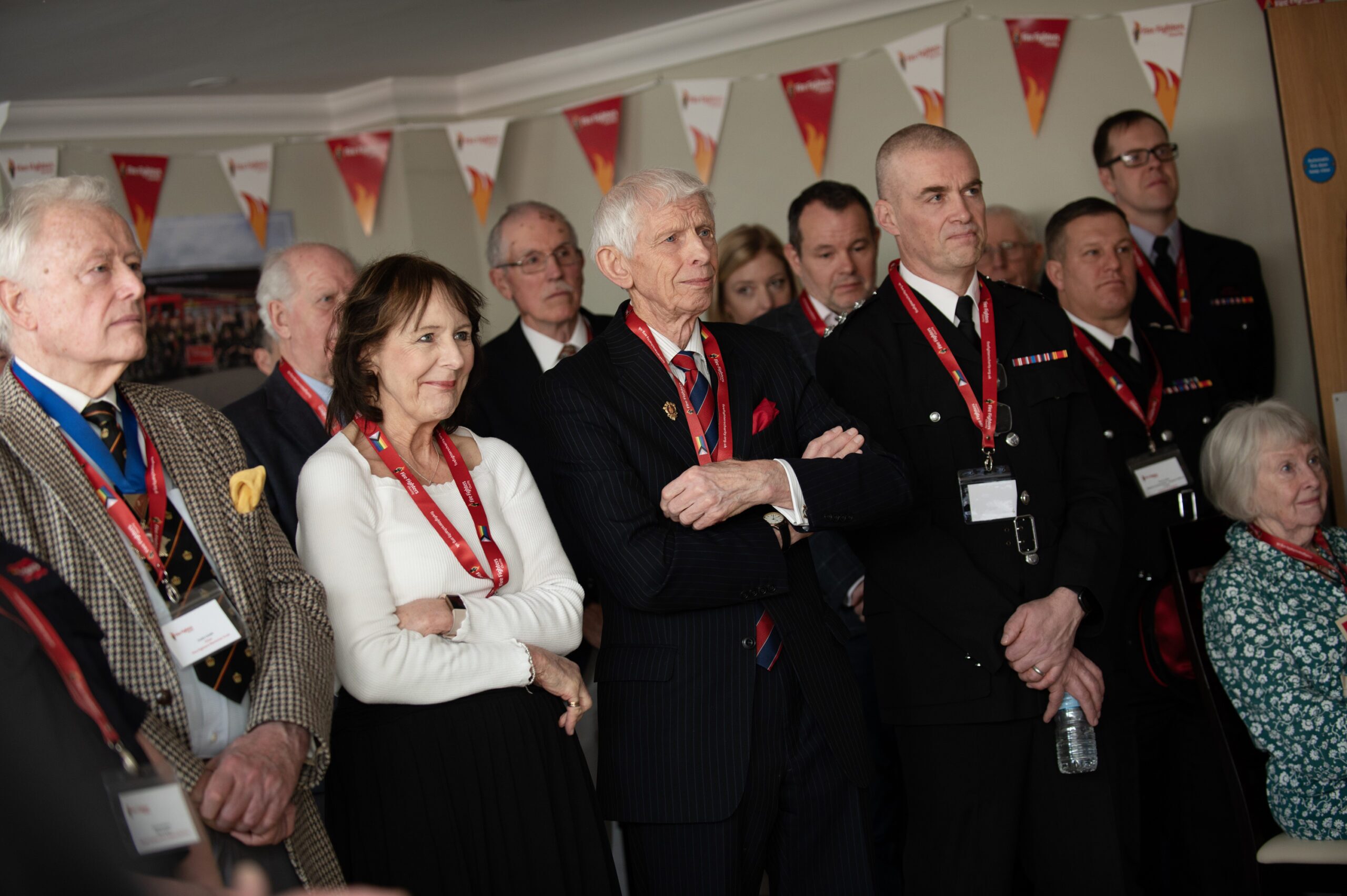 A group of people, mostly older adults, stand closely together indoors, wearing formal or business attire with red lanyards and name badges. Flags hang above them as they listen attentively to someone out of frame.