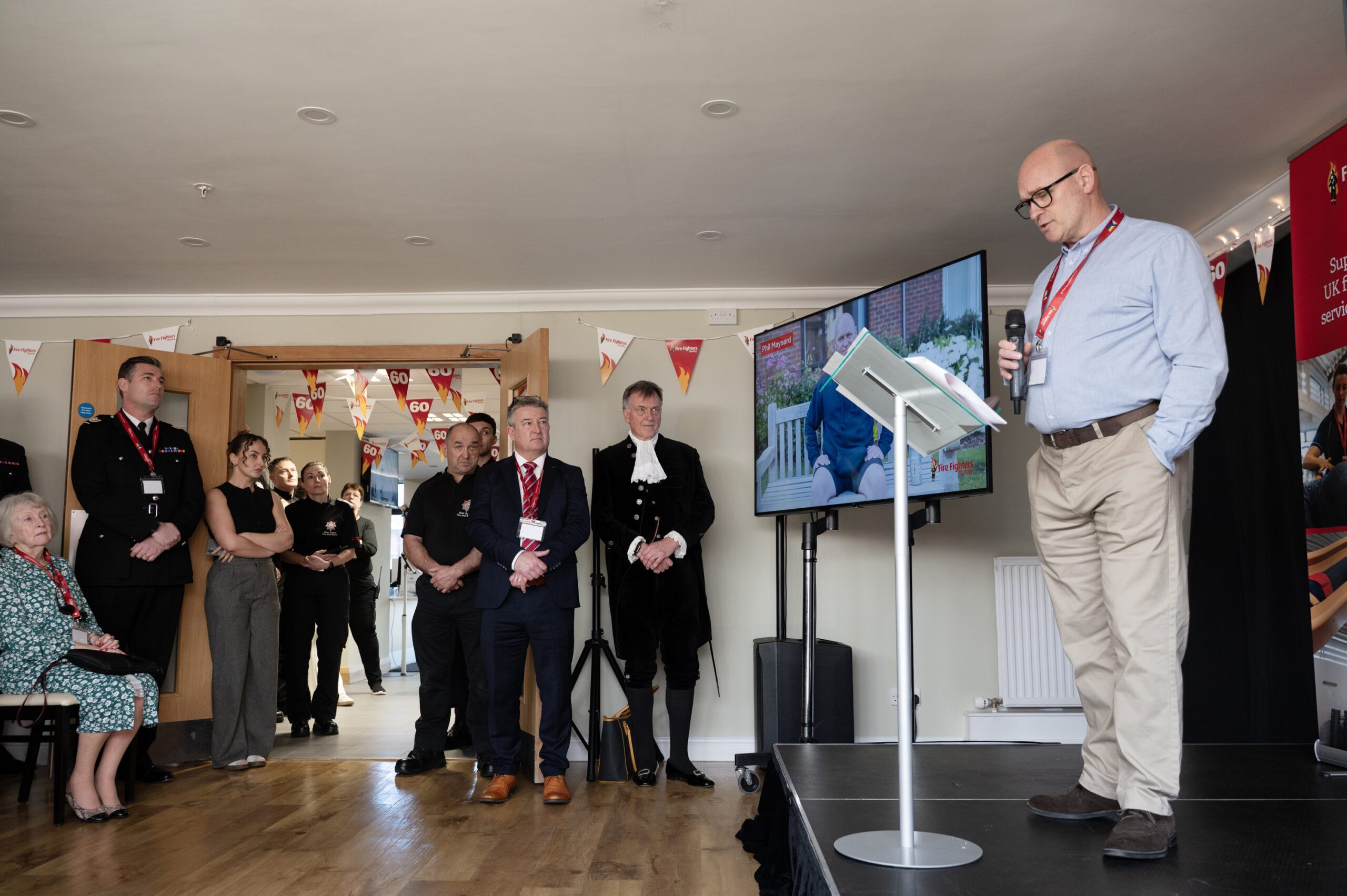 A man speaks at a podium on a small stage, addressing a group of people standing and listening in a decorated room. Some attendees wear uniforms, and a screen displays an image behind the speaker.