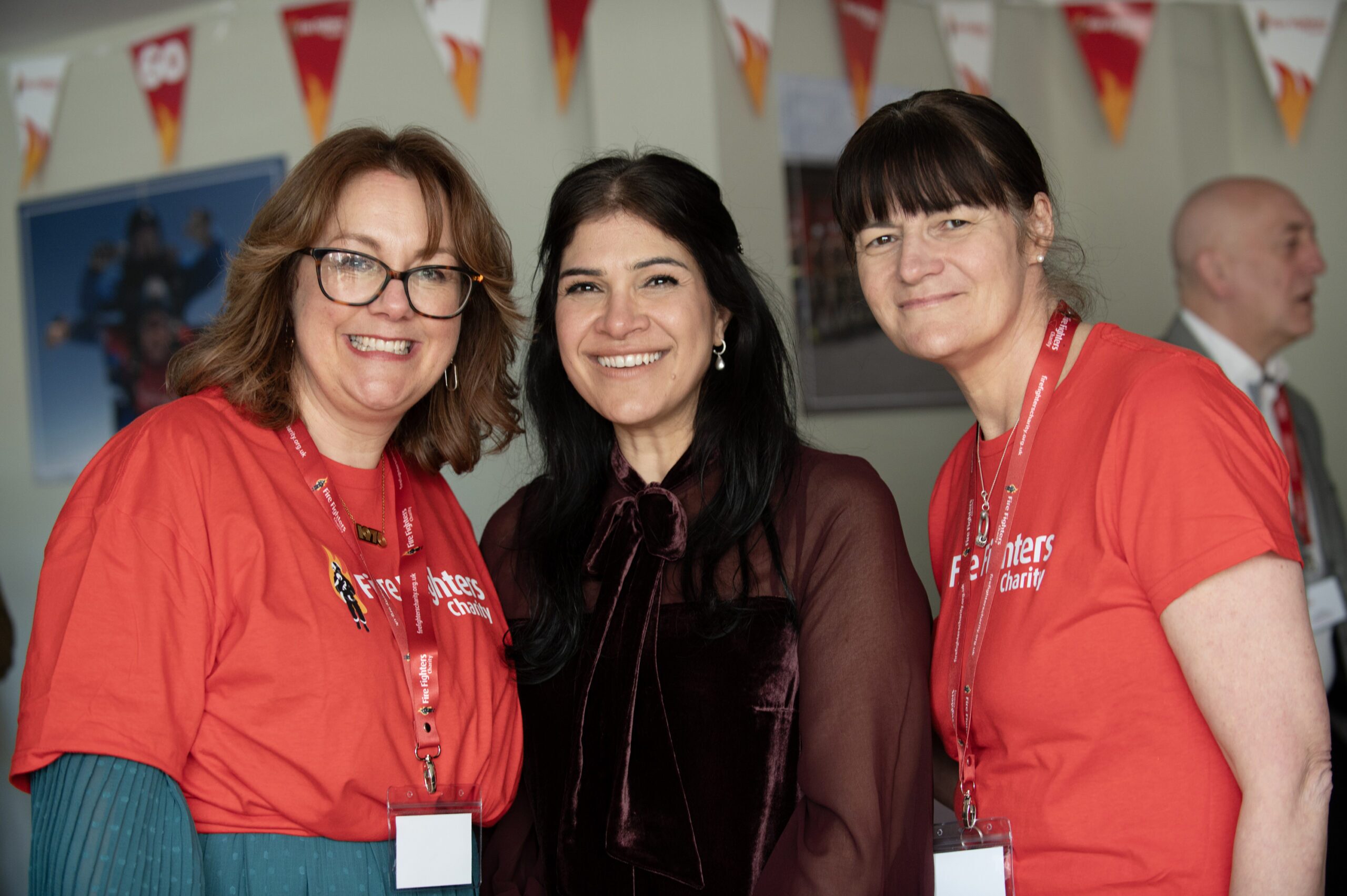 Three women smiling at the camera; two are wearing red shirts with lanyards and name badges, while the third wears a dark blouse. There are colorful triangular banners hanging in the background.