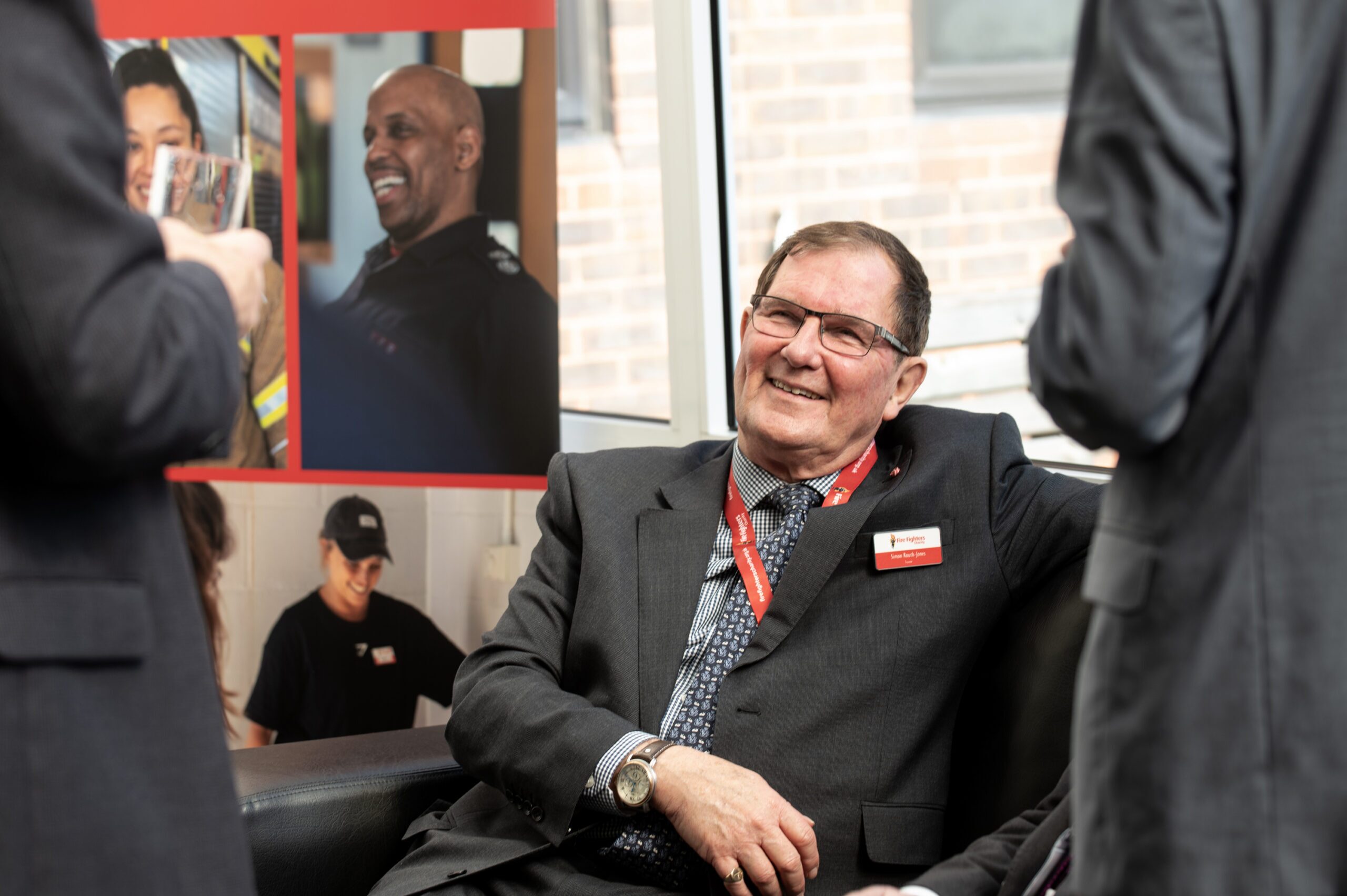 An older man wearing a suit and red lanyard smiles while sitting on a couch, engaging with others in conversation. In the background are posters with images of people and a window showing a brick building.