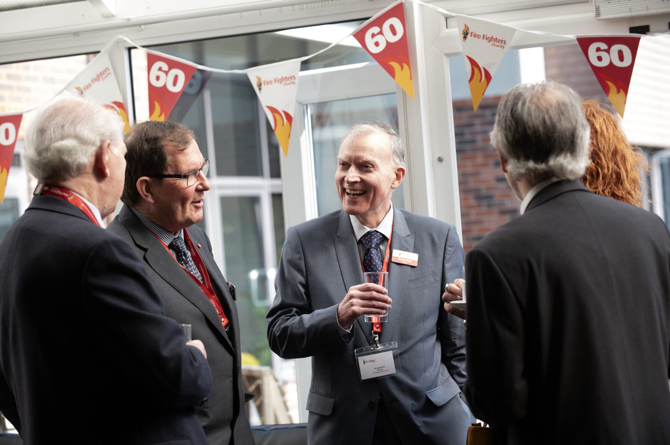 Five people in suits are talking and smiling at an indoor event with “60” and flame bunting decorations hanging above them. The atmosphere appears celebratory and professional.