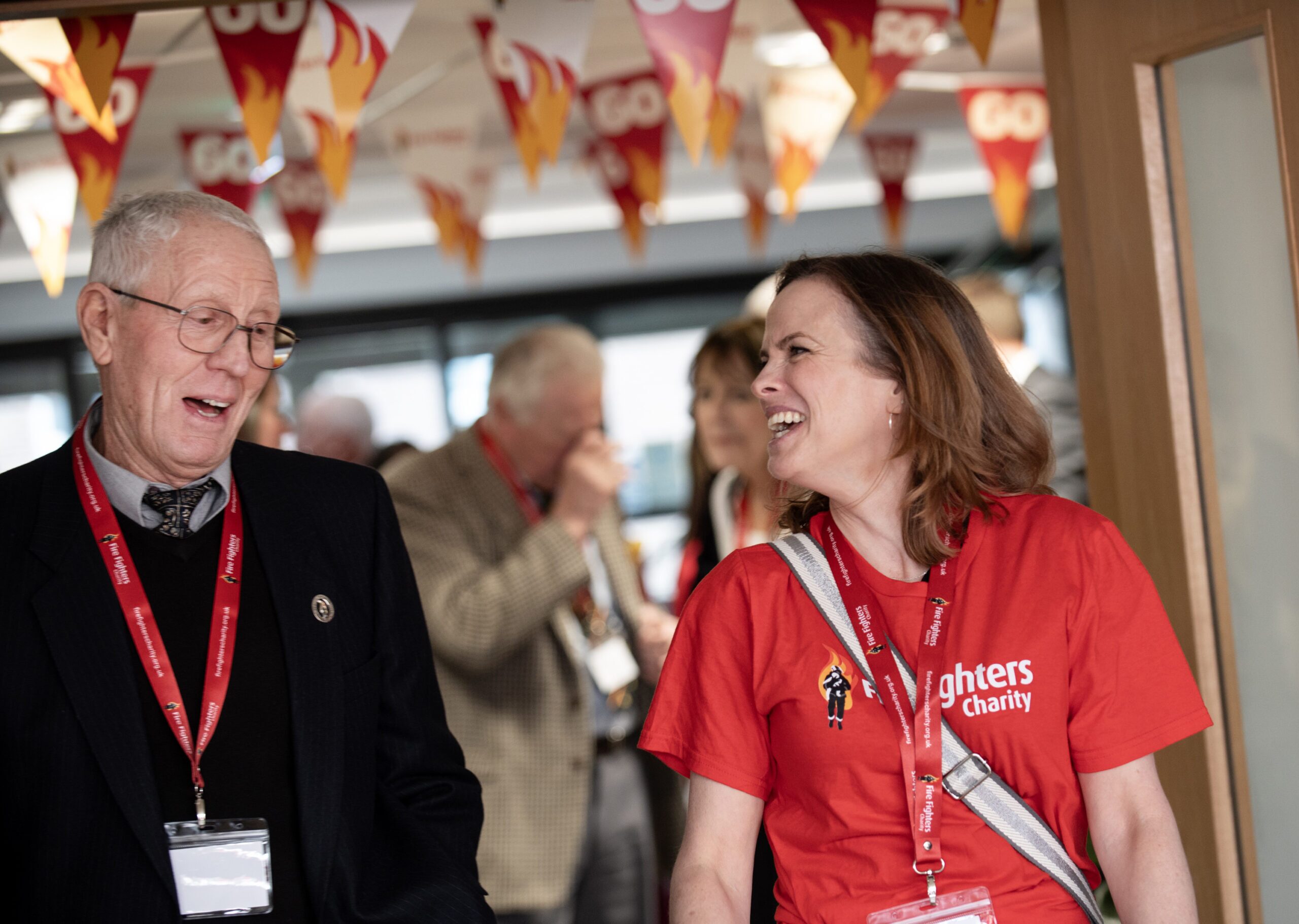 A smiling woman in a red charity t-shirt and an elderly man in a suit converse at an indoor event decorated with red and yellow bunting, while other people mingle in the background.