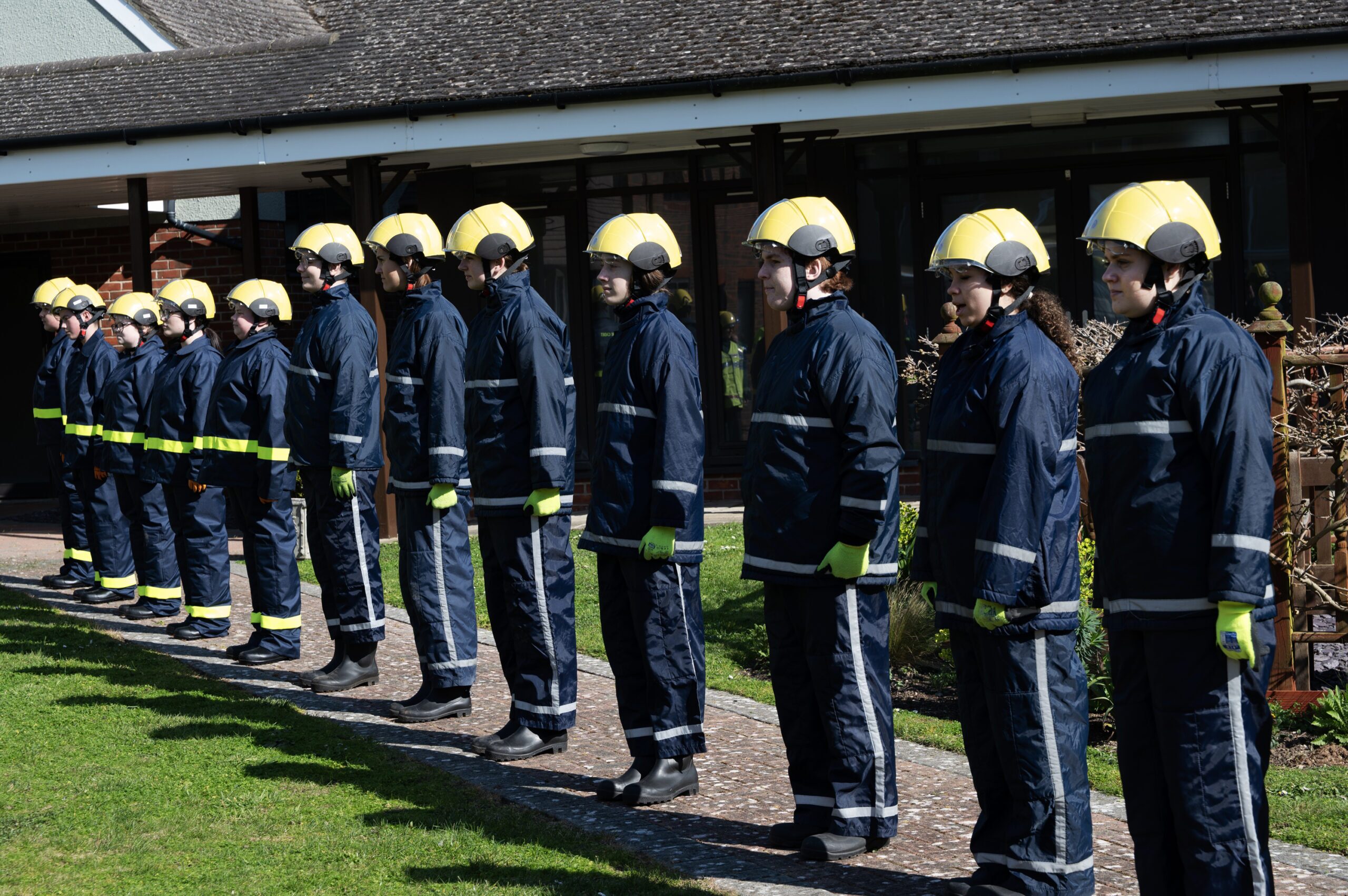 A group of firefighters in uniform and yellow helmets stand in a straight line outdoors, facing forward, near a building with large windows.