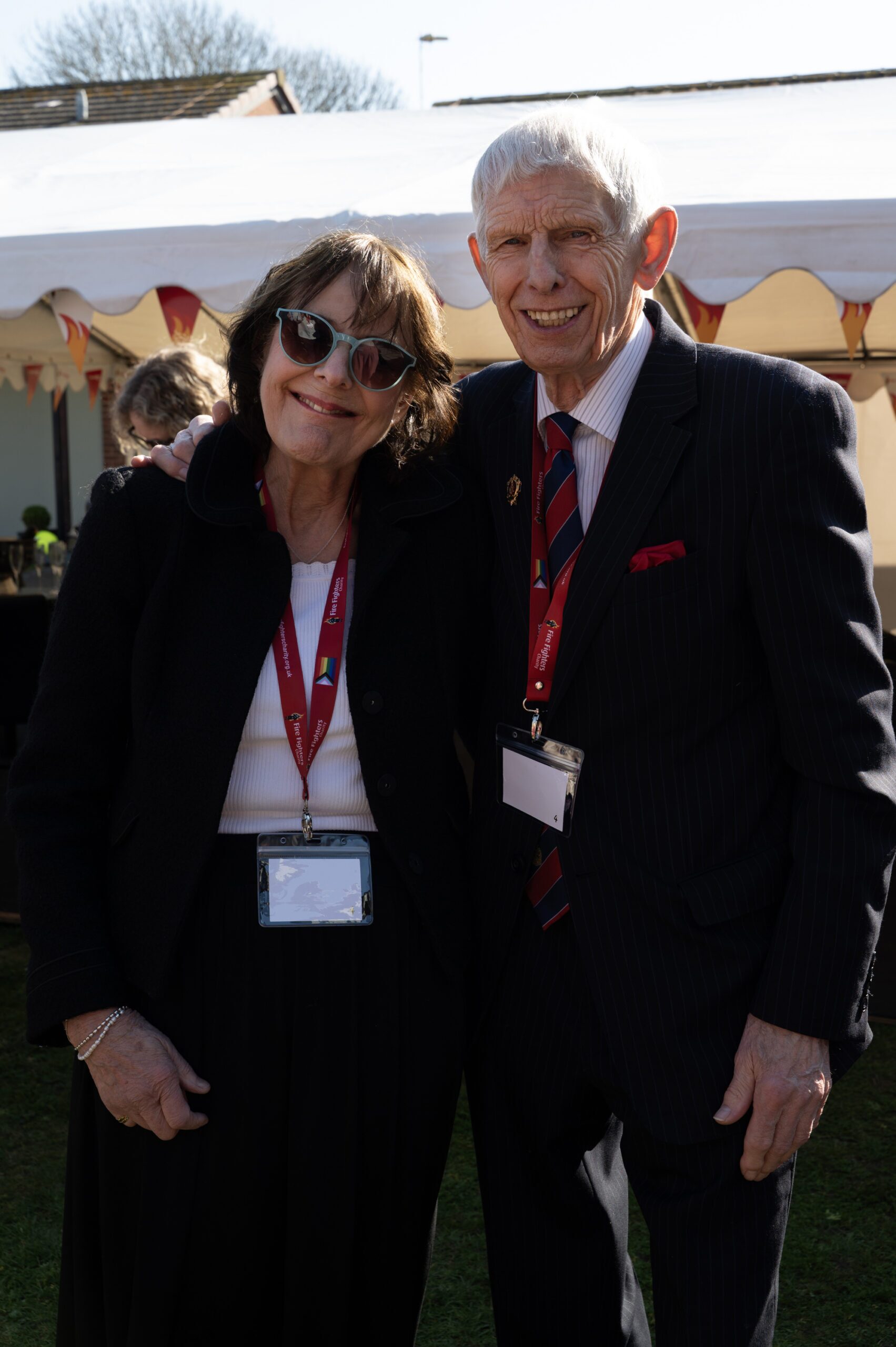 Two older adults stand close together outdoors, smiling at the camera. Both wear name badges and red lanyards; the woman has sunglasses and dark hair, and the man wears a suit and striped tie. A tent is visible behind them.