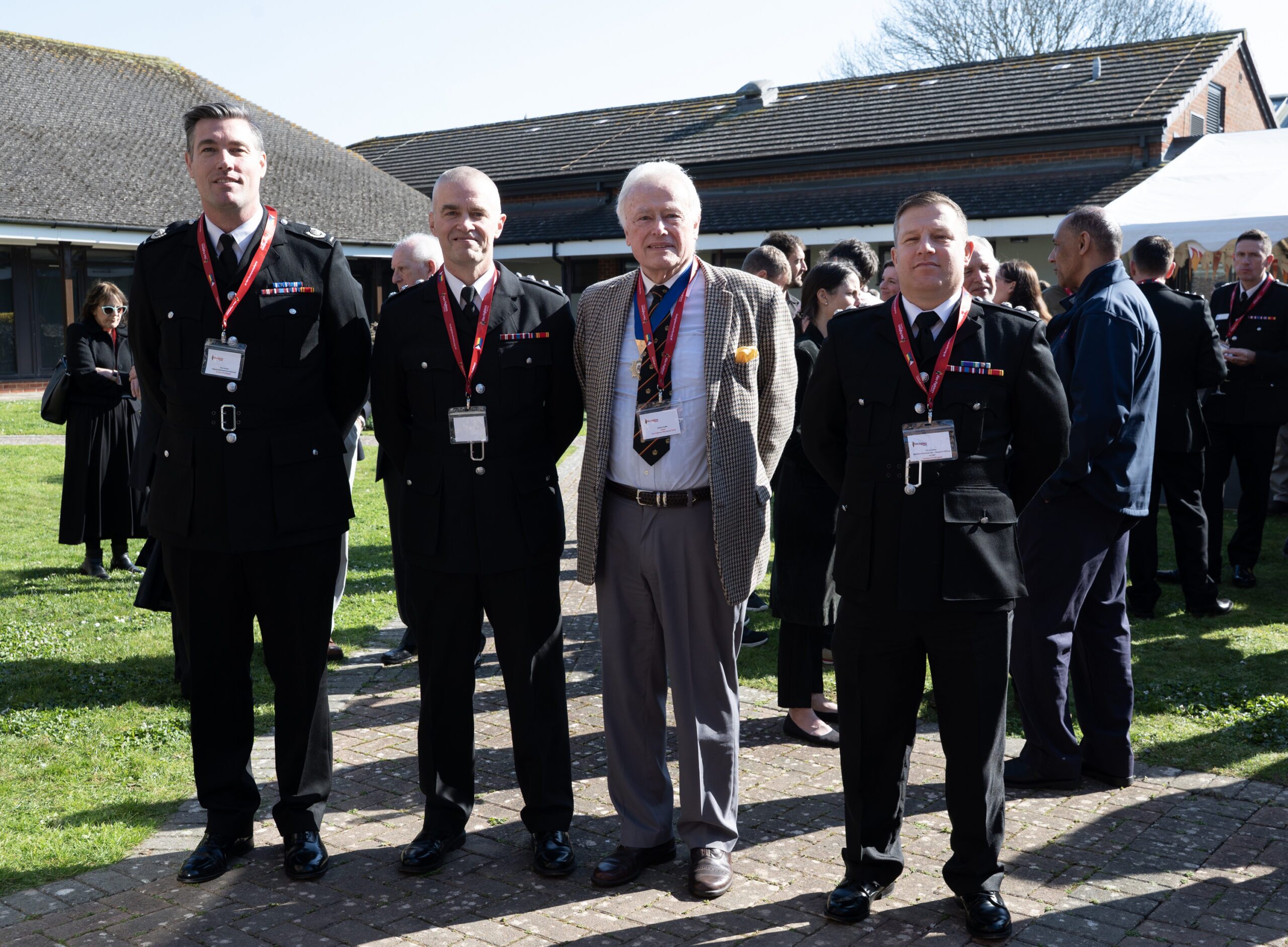 Four men stand outside in a sunny courtyard, three in formal black uniforms with medals and one in a light plaid suit. Other people are gathered in the background, near brick buildings and green grass.