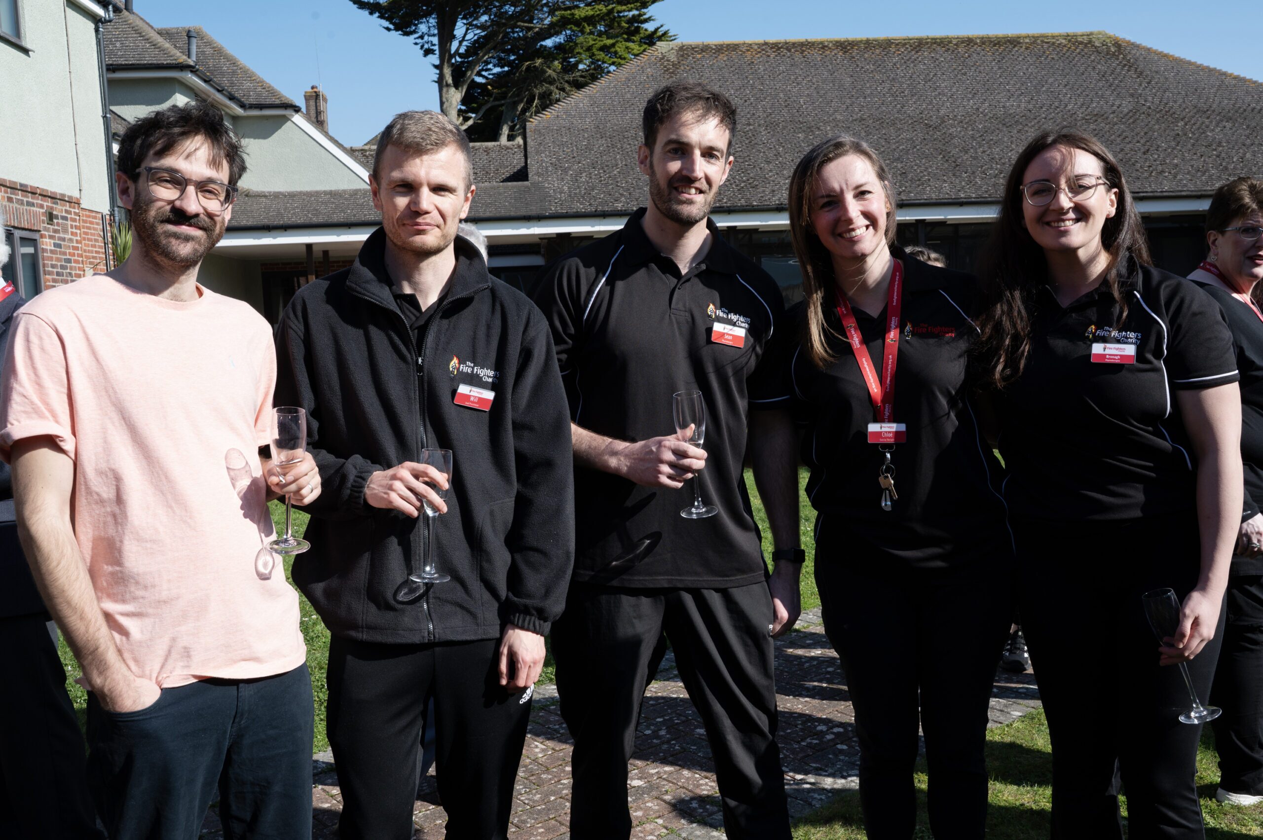 Five people stand outside, smiling at the camera. Three wear black shirts with logos, one wears a light pink shirt, and one a black top with a red lanyard. Some hold empty champagne flutes. It’s sunny, with buildings in the background.