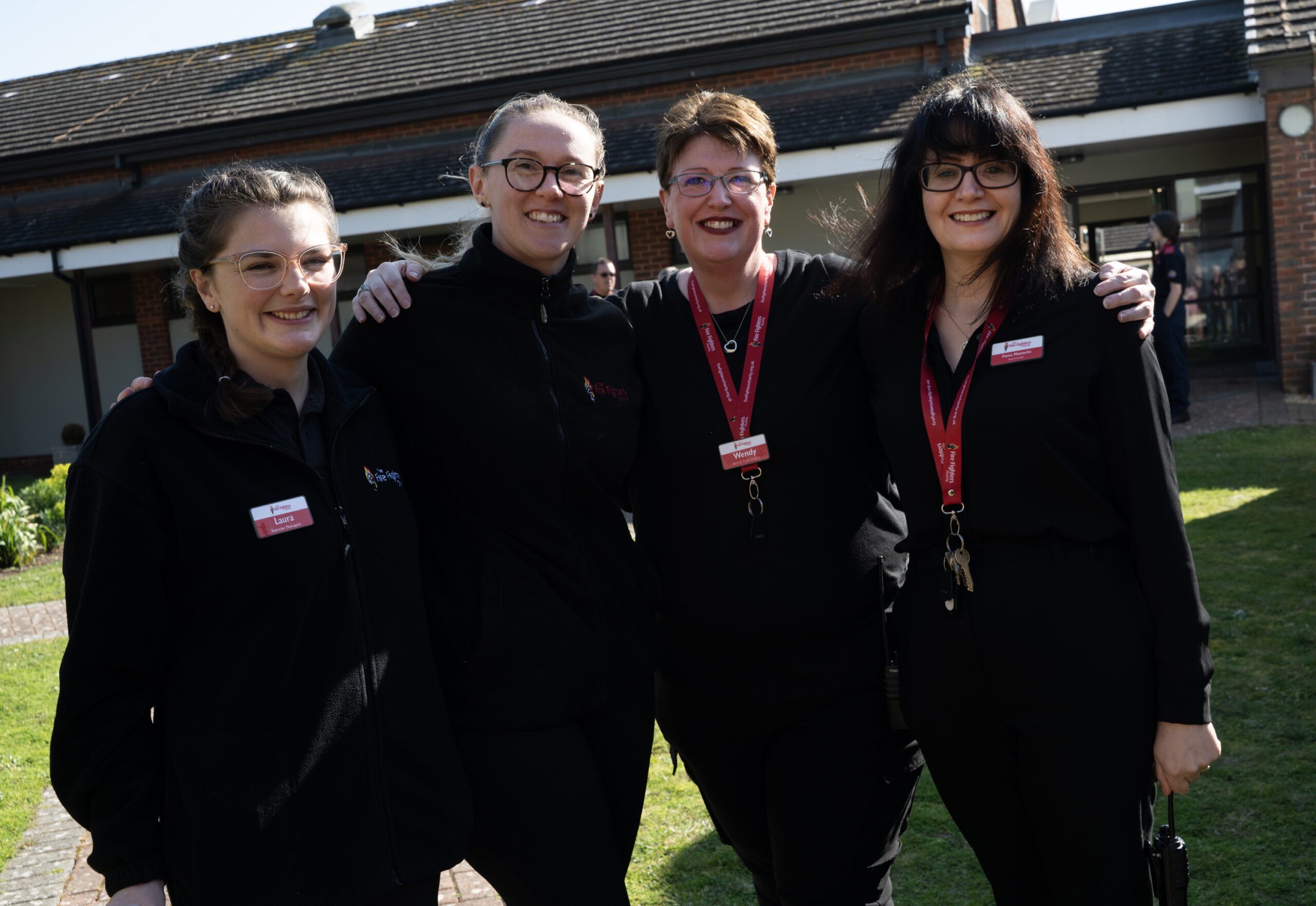 Four smiling women wearing black outfits and name badges stand close together outdoors in front of a building, posing for a group photo on a sunny day.