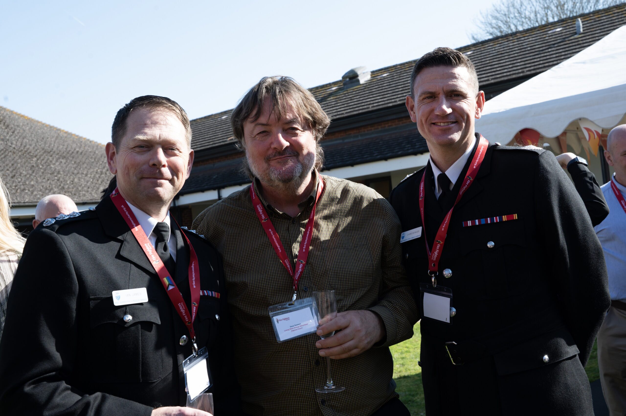 Three men pose outdoors at a daytime event. Two wear black formal uniforms with medals and red lanyards; the man in the center wears a brown shirt, red lanyard, and holds a glass. People and tents are visible in the background.