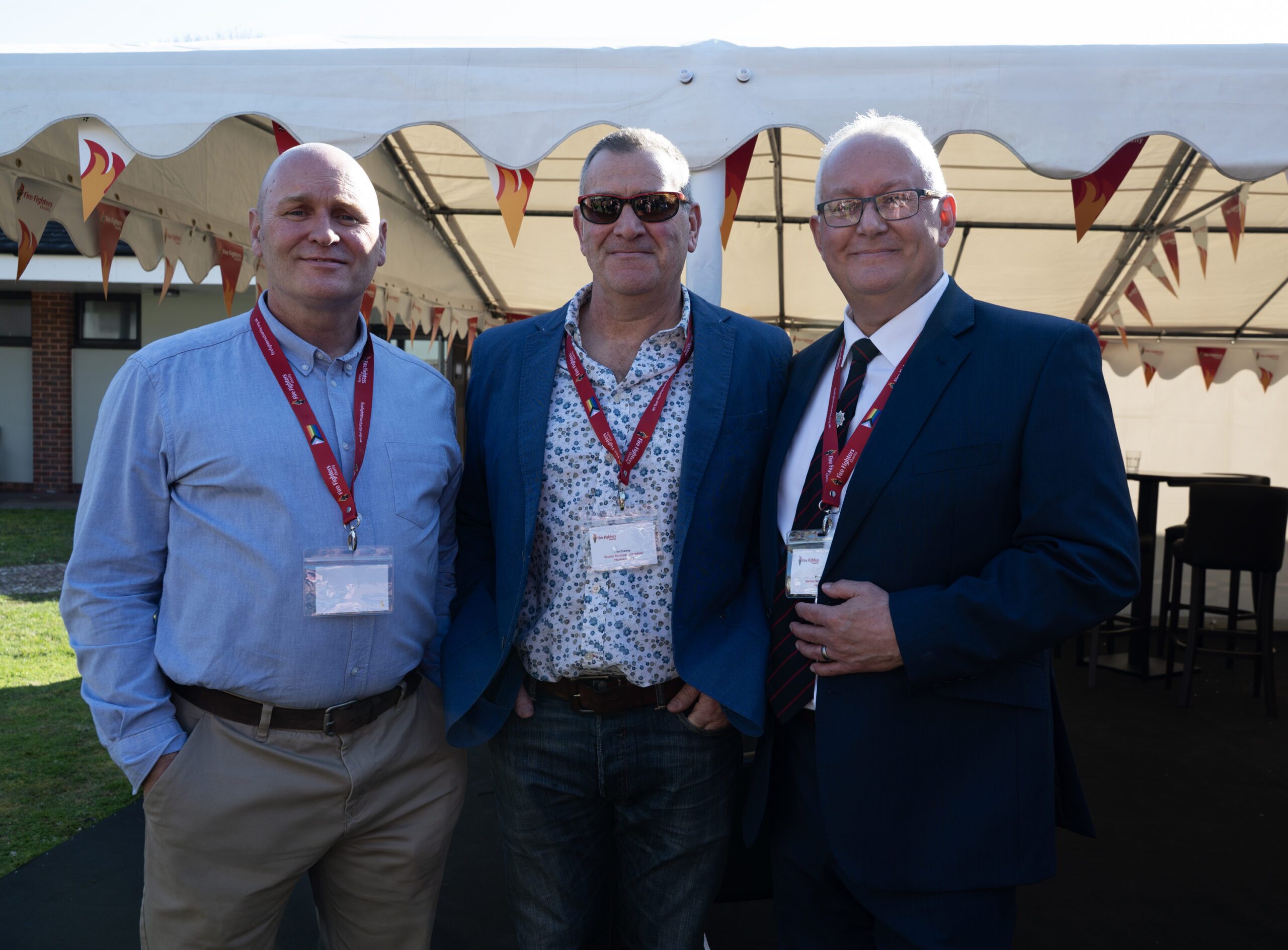 Three men wearing name badges and red lanyards stand together under a white outdoor tent. Two are in business attire, one wears a casual shirt. They are smiling and posing for the camera at an event.