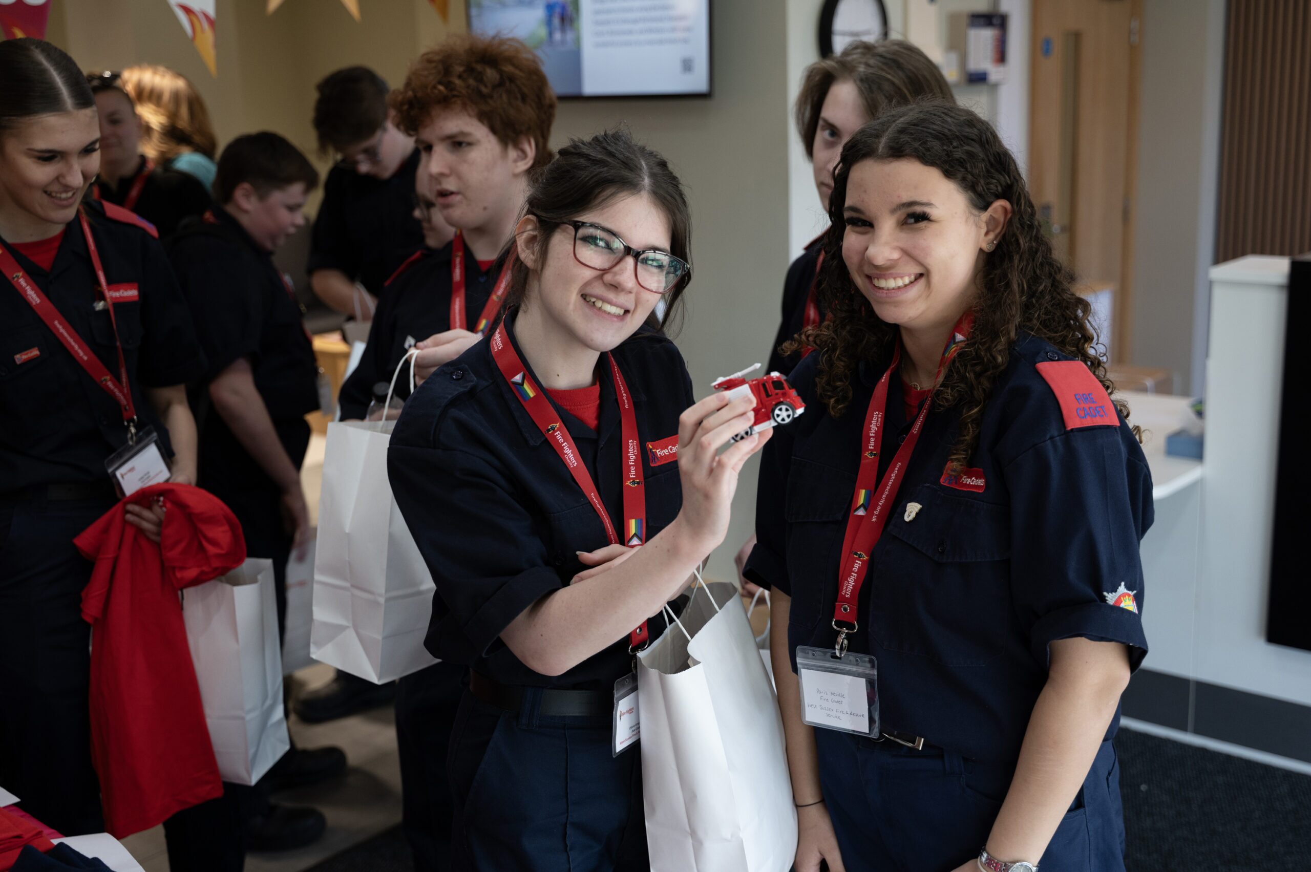 A group of young people in dark uniforms with red lanyards stand together indoors. Two of them smile at the camera, one holding a toy fire truck. They each have a white gift bag. Other people are chatting in the background.