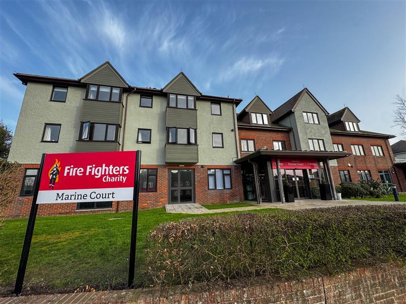 A large building with a sign in front that reads "Fire Fighters Charity Marine Court." The building has brick and light green walls with multiple windows and a clear blue sky overhead.