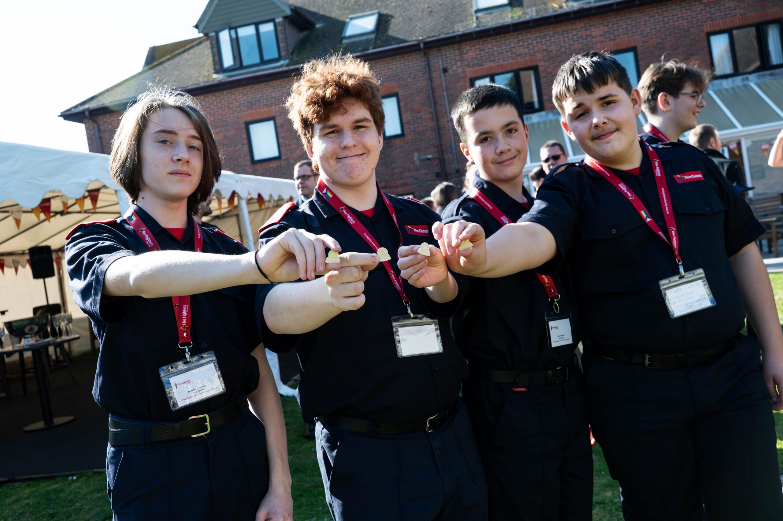 Four teenagers in matching uniforms and red lanyards smile and hold a wooden stick together outdoors, with a brick building and a white tent in the background.