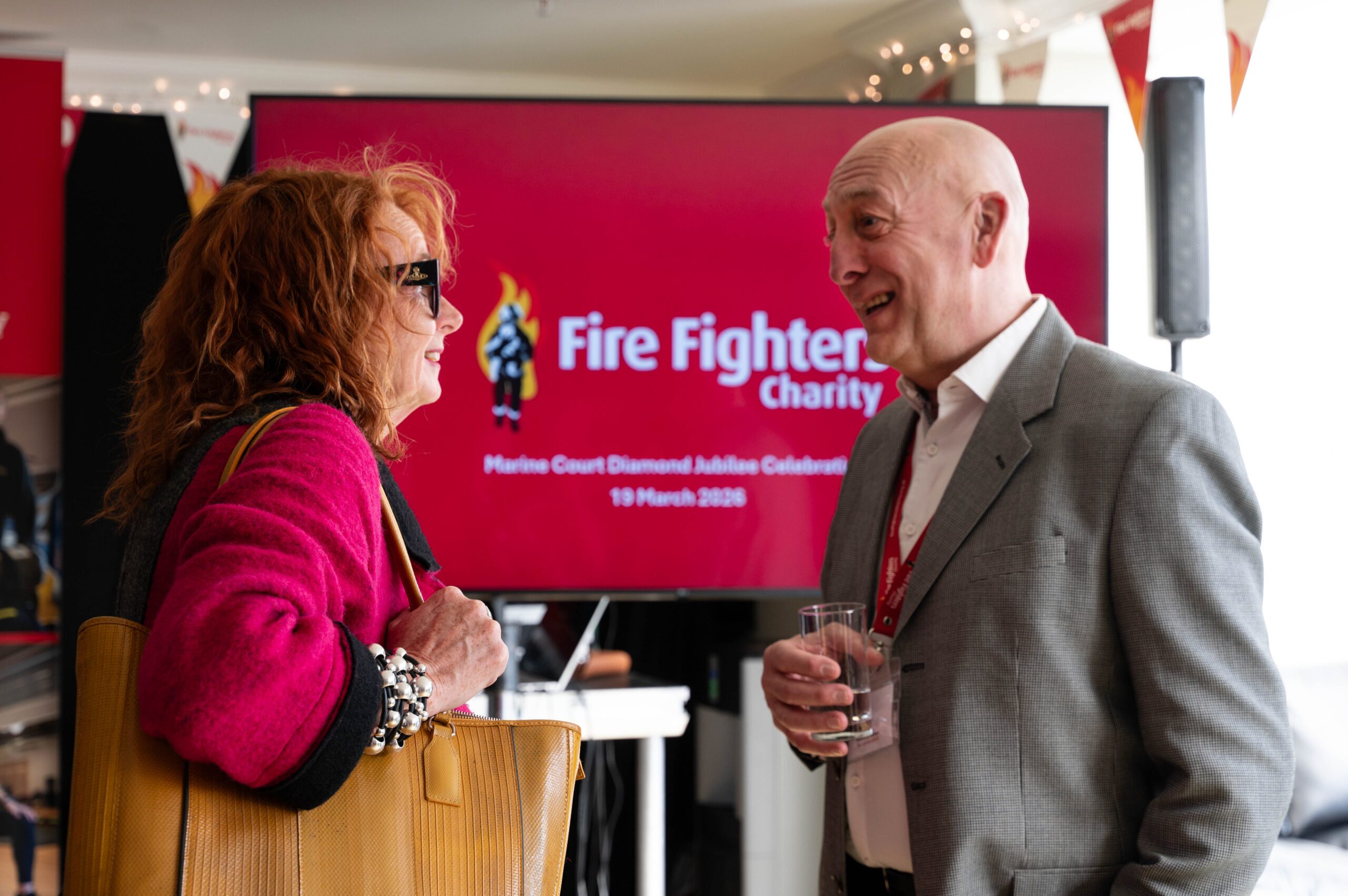A woman in a bright pink coat with a large tan bag talks to a smiling man in a gray suit holding a glass. Behind them is a screen displaying "Fire Fighters Charity" at an indoor event.