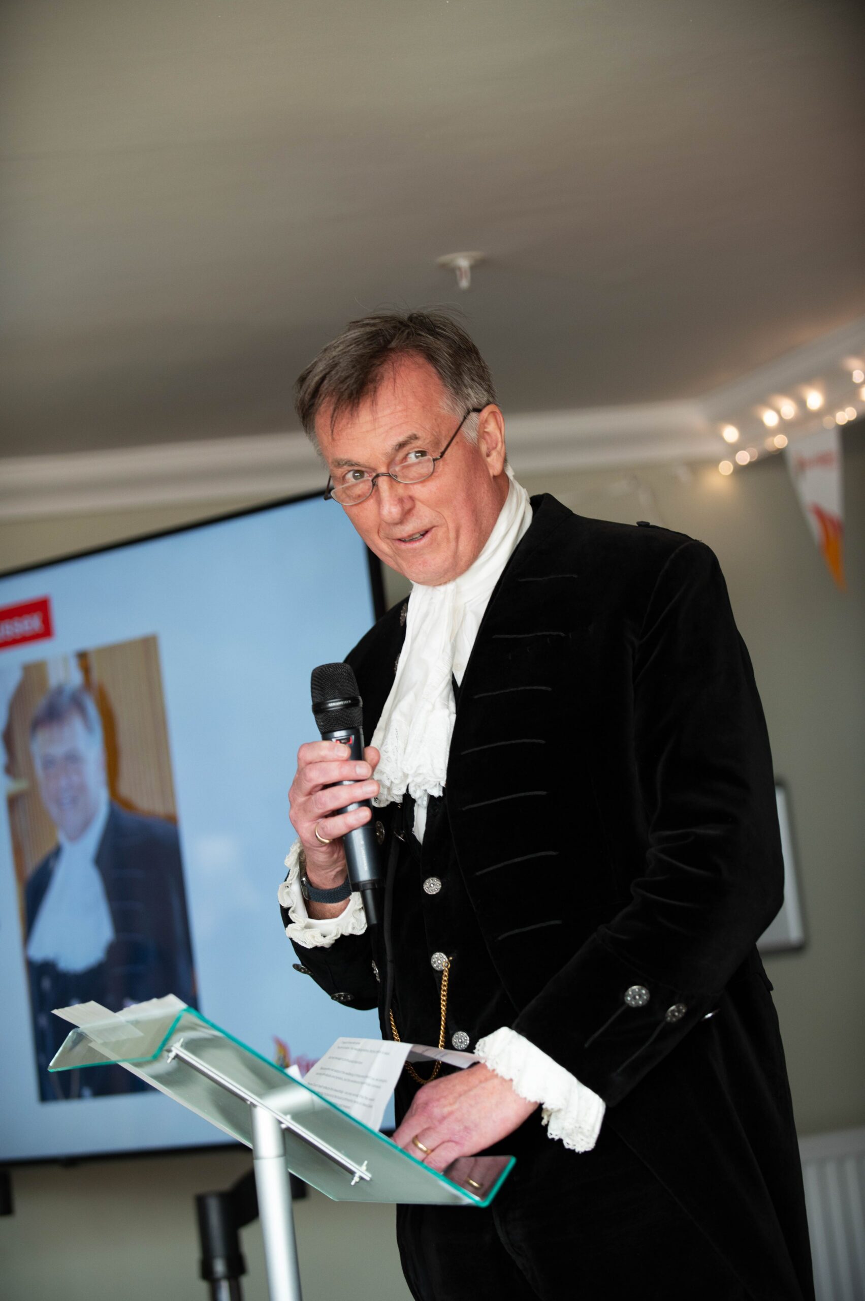 A man in formal, historical clothing speaks into a microphone while standing at a podium. A screen behind him displays a photo of the same man in similar attire. The setting appears to be indoors with soft lighting.