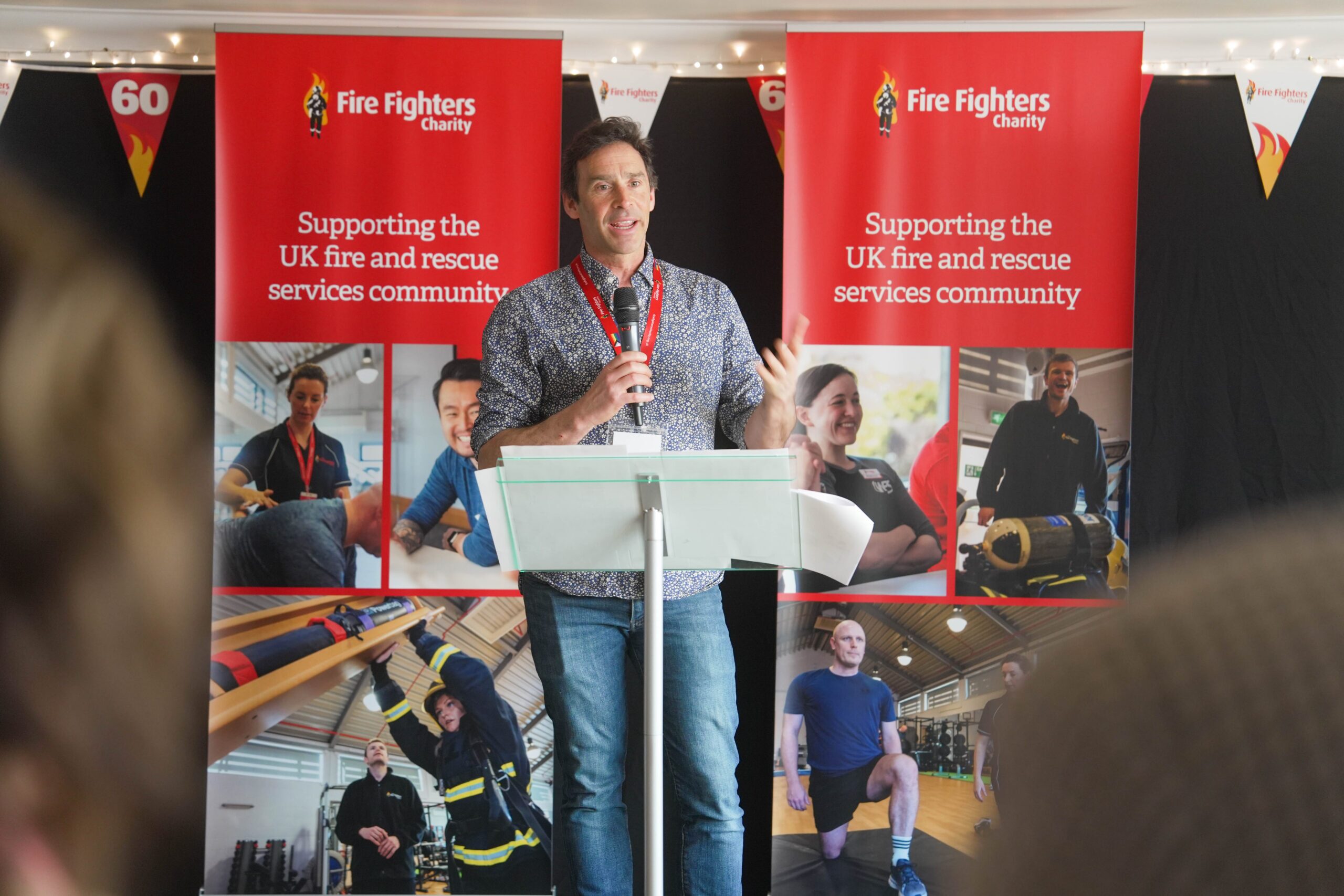 A man speaks at a podium in front of banners for the Fire Fighters Charity, which support the UK fire and rescue services community. People are visible in photos on the banners behind him.