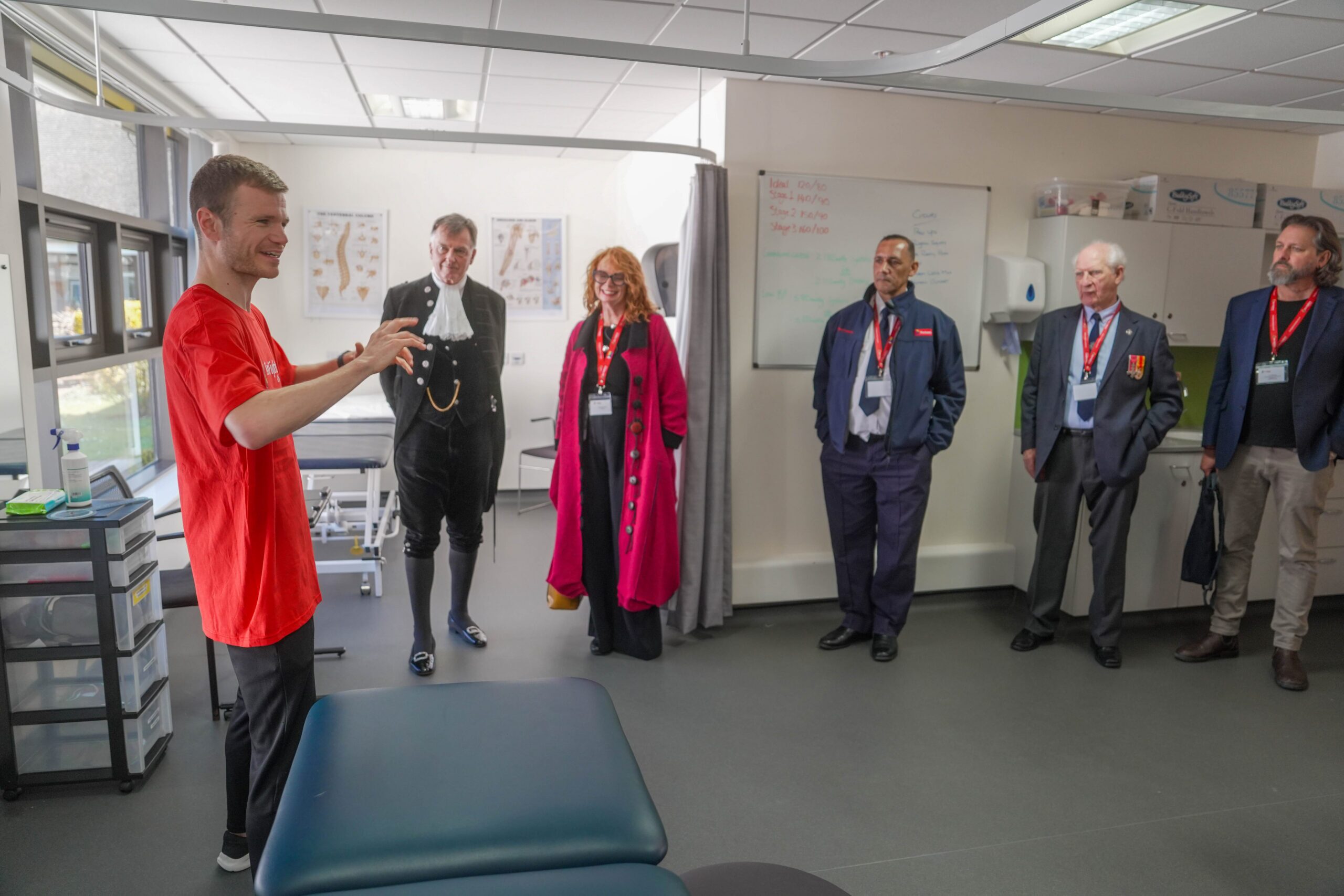 A man in a red shirt gestures while speaking to five adults standing in a medical examination room; some wear formal attire and name badges, and anatomy charts are visible on the wall behind them.